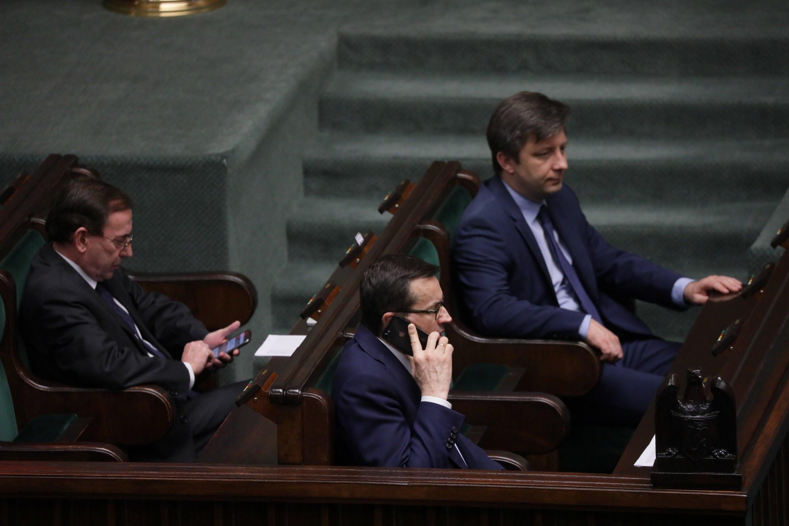 Mateusz Morawiecki rozmawia przez telefon w ławach rządowych na sali sejmowej. Na drugim planie Michał Dworczyk