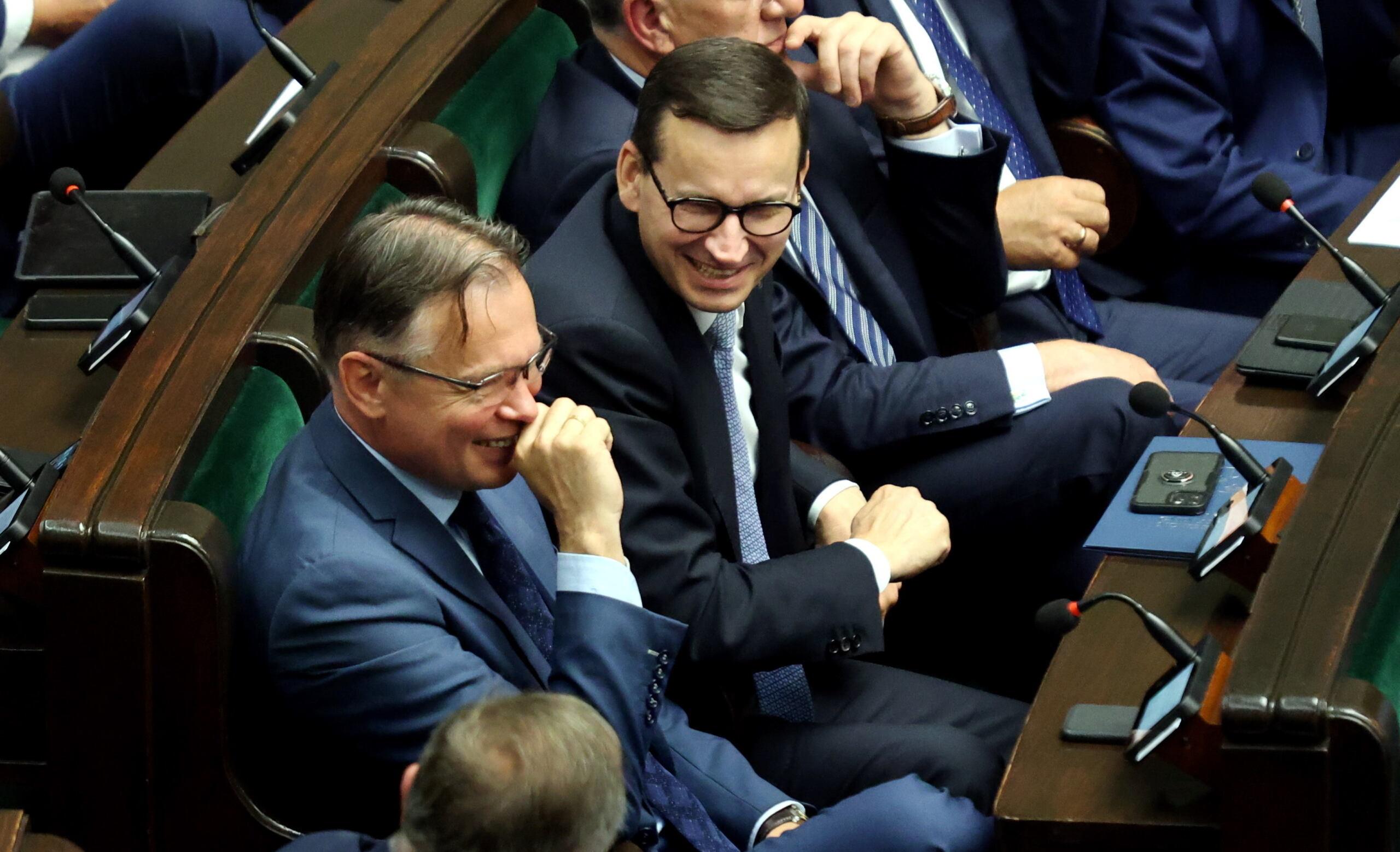 W ławach rządowych w Sejmie siedzi Arkadiusz Mularczyk śmiejący się, obok niego uśmiechnięty Mateusz Morawiecki