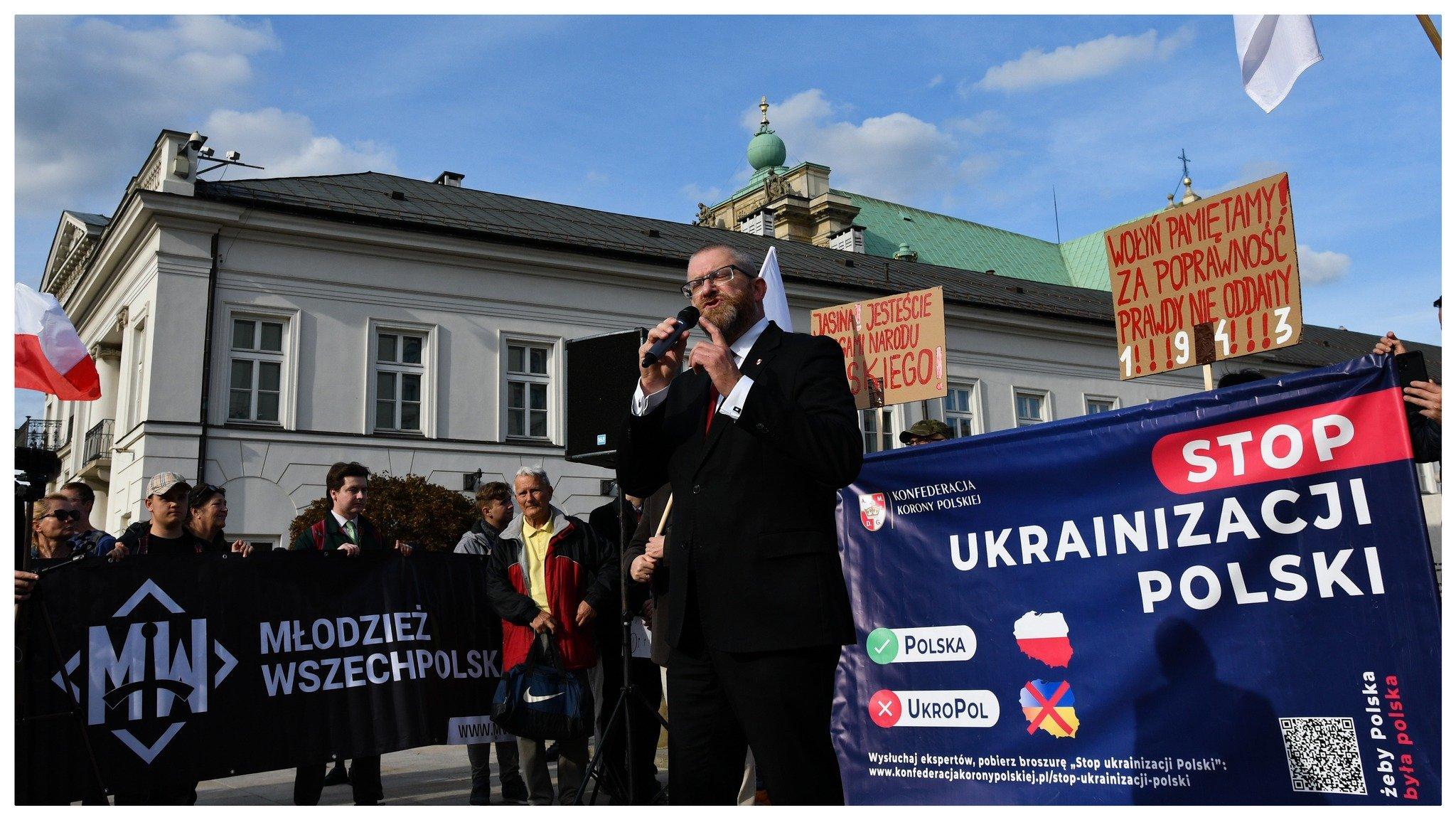 Poseł KOnfederacji Grzegorz Braun stoi z mikrofonem przed transparentem "Ukrainizacja Polski", obok inni ludzie