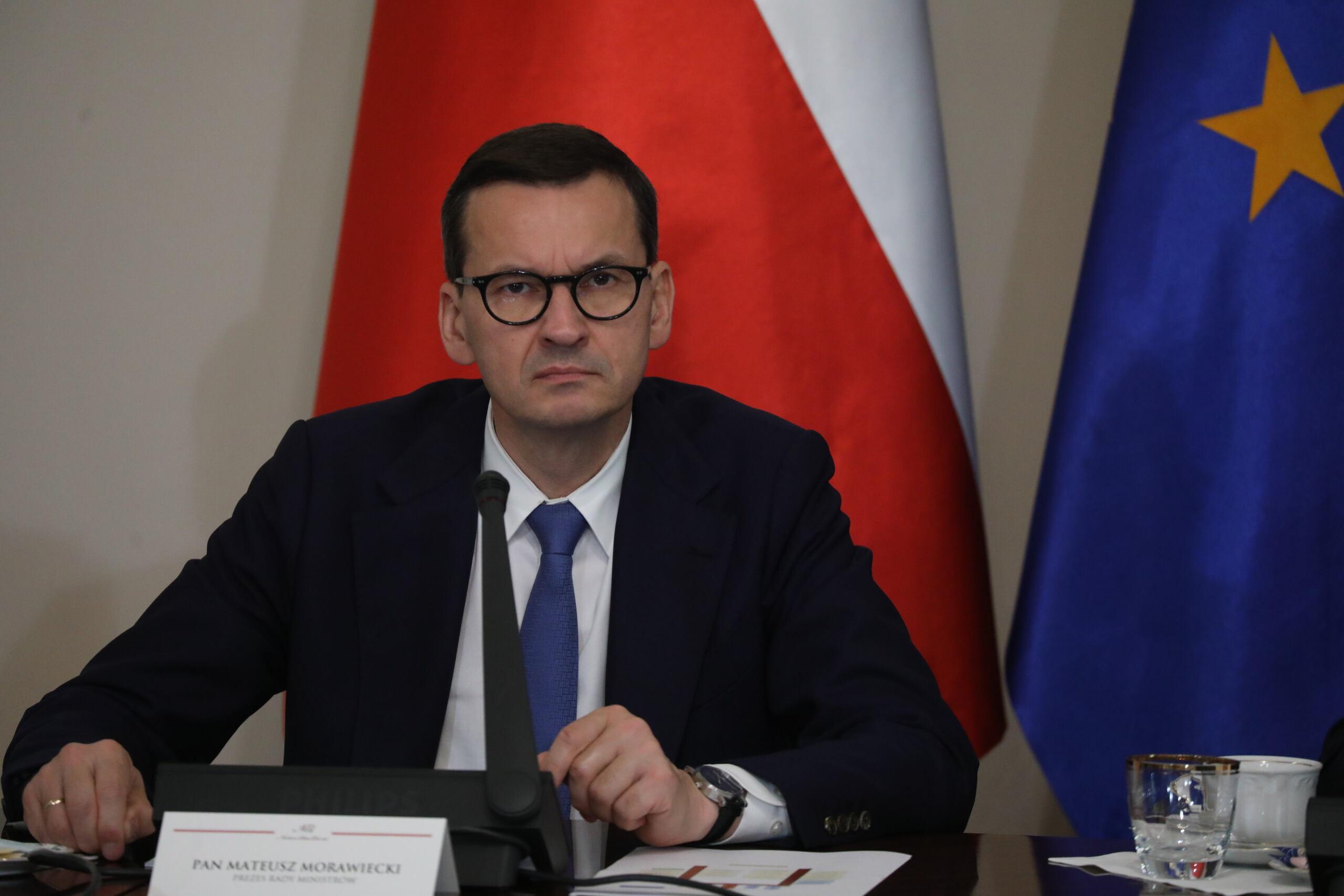 Premier Morawiecki z ponurą miną patrzy w obiektyw. Za nim flagi Polski i Unii Europejskiej