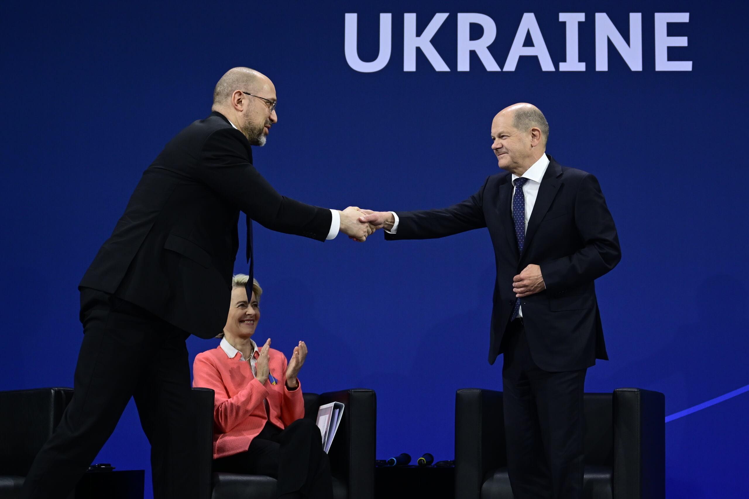 Premier Ukrainy na scenie śsciska rękę kanclerza Nioemiec, w tele siedzi Ursula von der Leyen