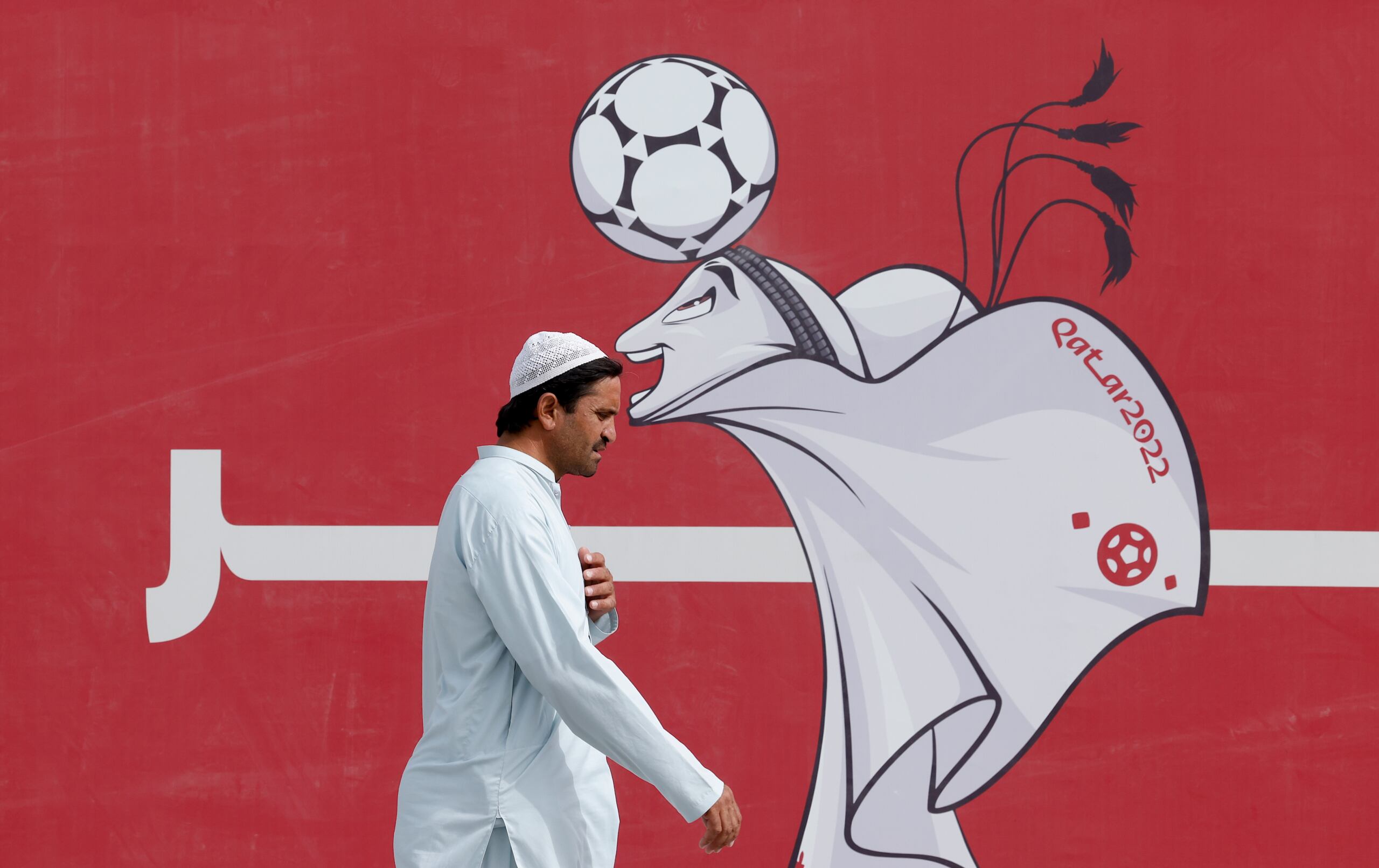 Mężczyzna w białej szacie przechodzi obok wizerunku maskotki mundialu w Katarze, namalowanej na czerwonym tle