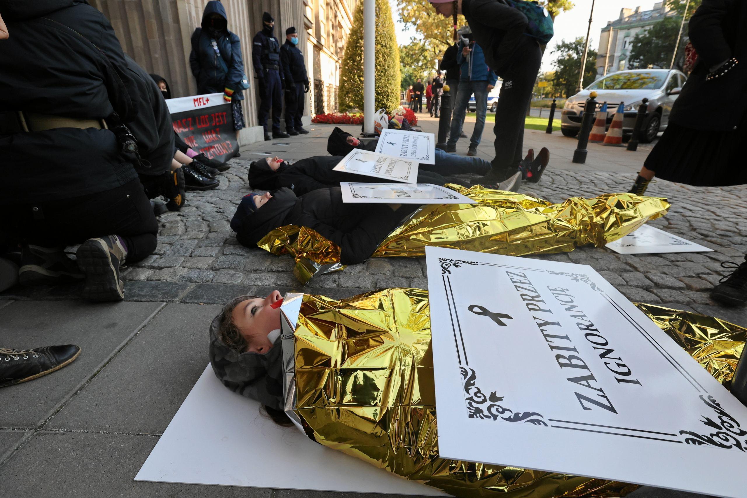 Ludzie zawinięci w koce termiczne leżą na chodniku z symbolicznymi klepsydrami mówiącymi, ze sa ofiarami zniszczonej przez Czarnka edukacji