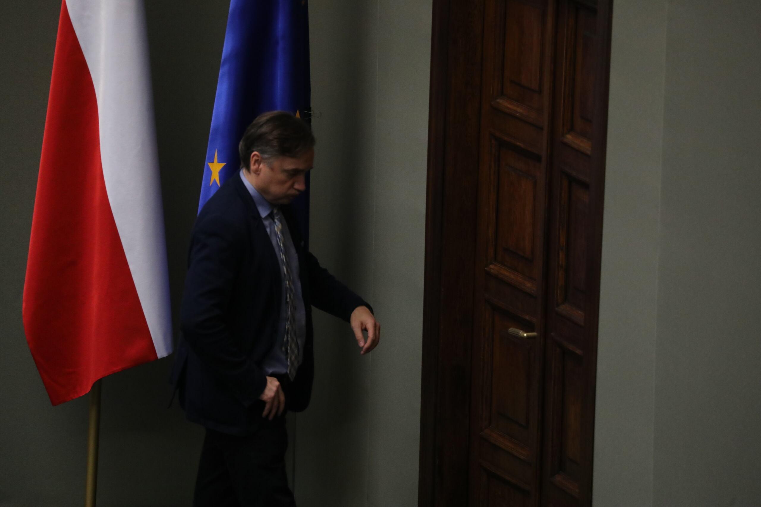 Zbigniew Ziobro podchodzi do drzwi z pochyloną głową. W tle flagi Polski i UE
