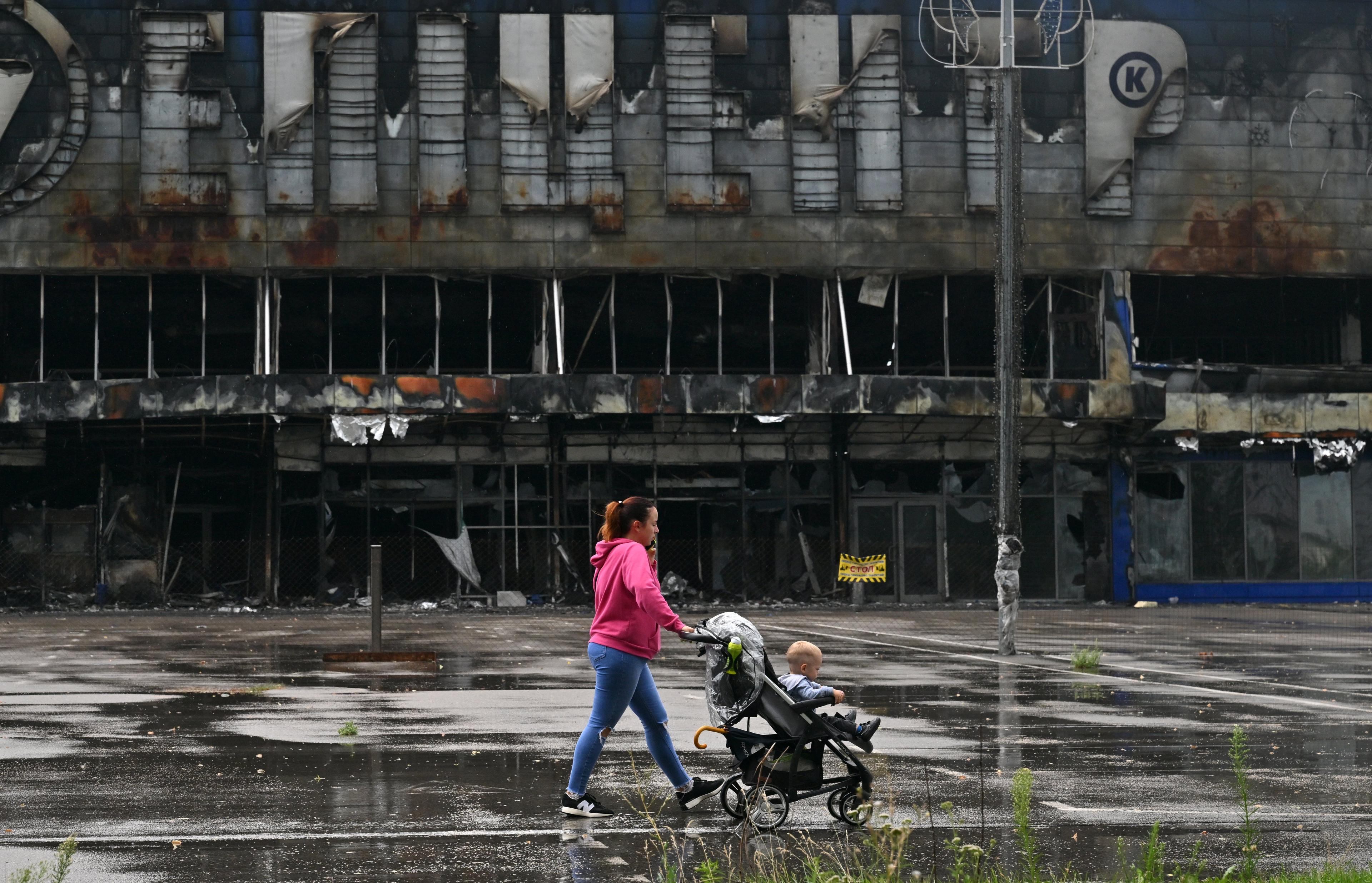 Kobieta z małym dzieckiem w wózku, w tle zniszczone centrum handlowe w Buczy