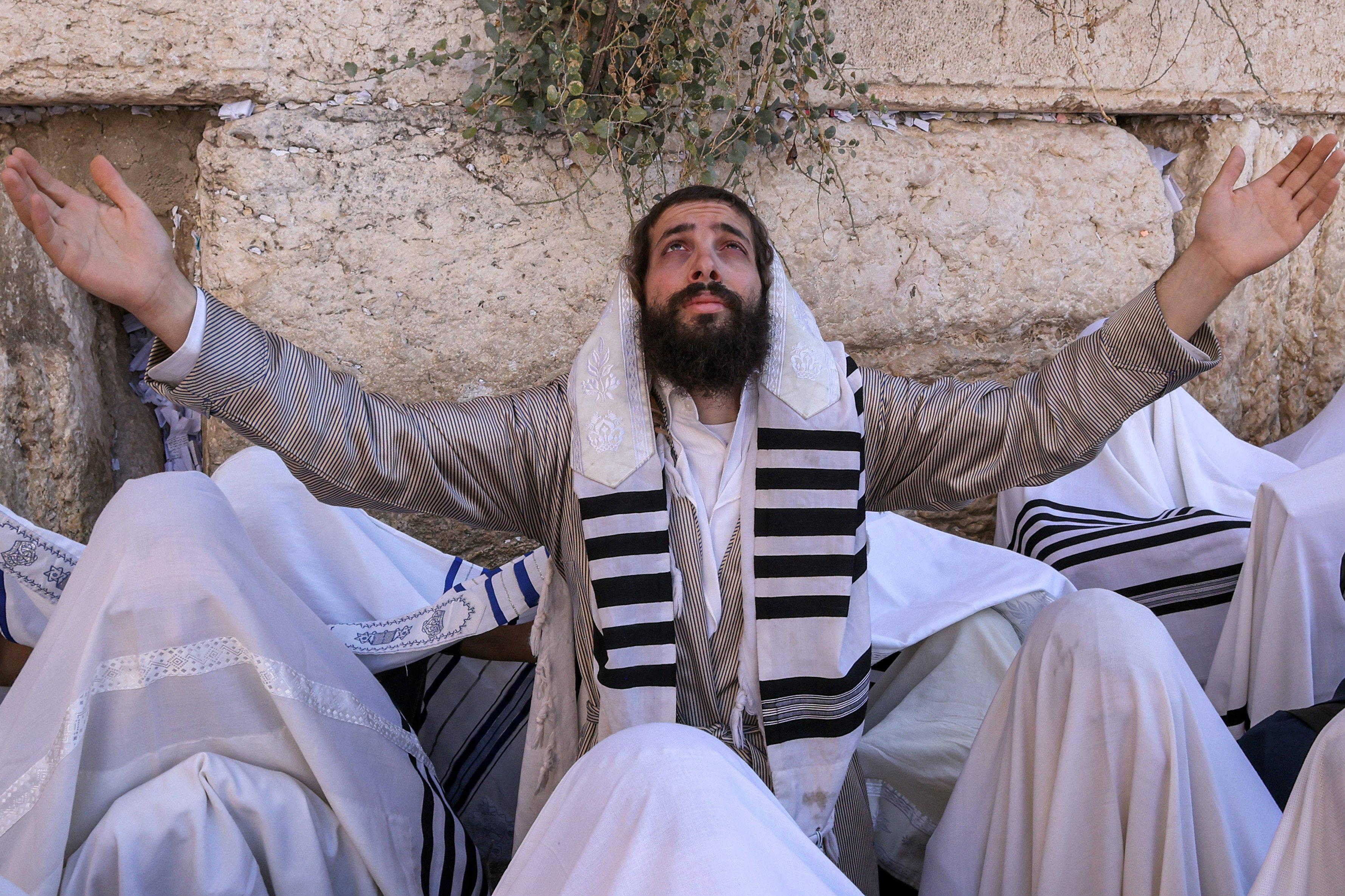 Żyd w ortodoksyjnym stroju, stojący wśrod innych Żydów ortodoksyjnych, podnosi ręce w geście modlitwy na tle muru