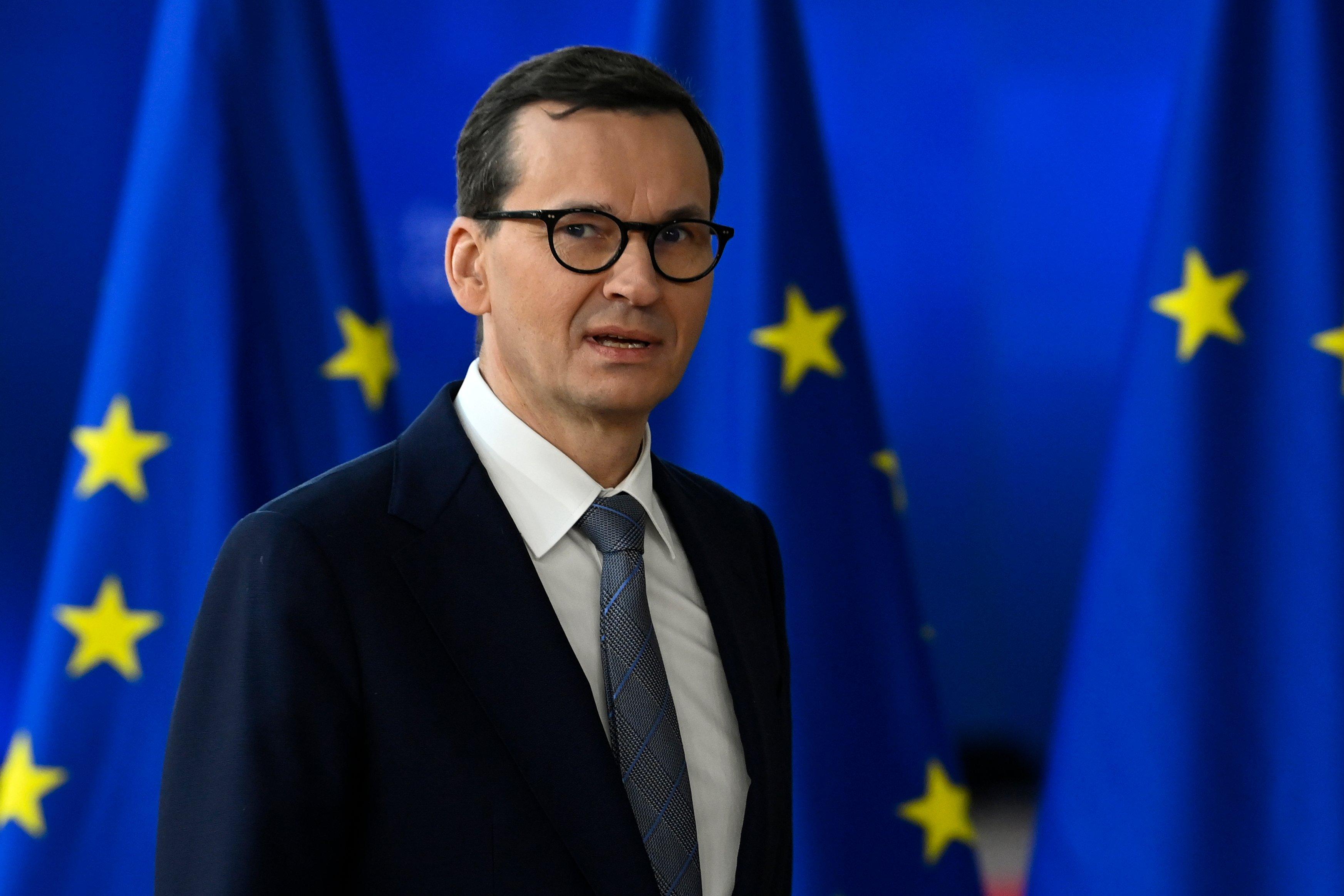 Mężczyzna w średnim wieku w okularach na tle flag Unii Europejskiej. To Mateusz Morawiecki