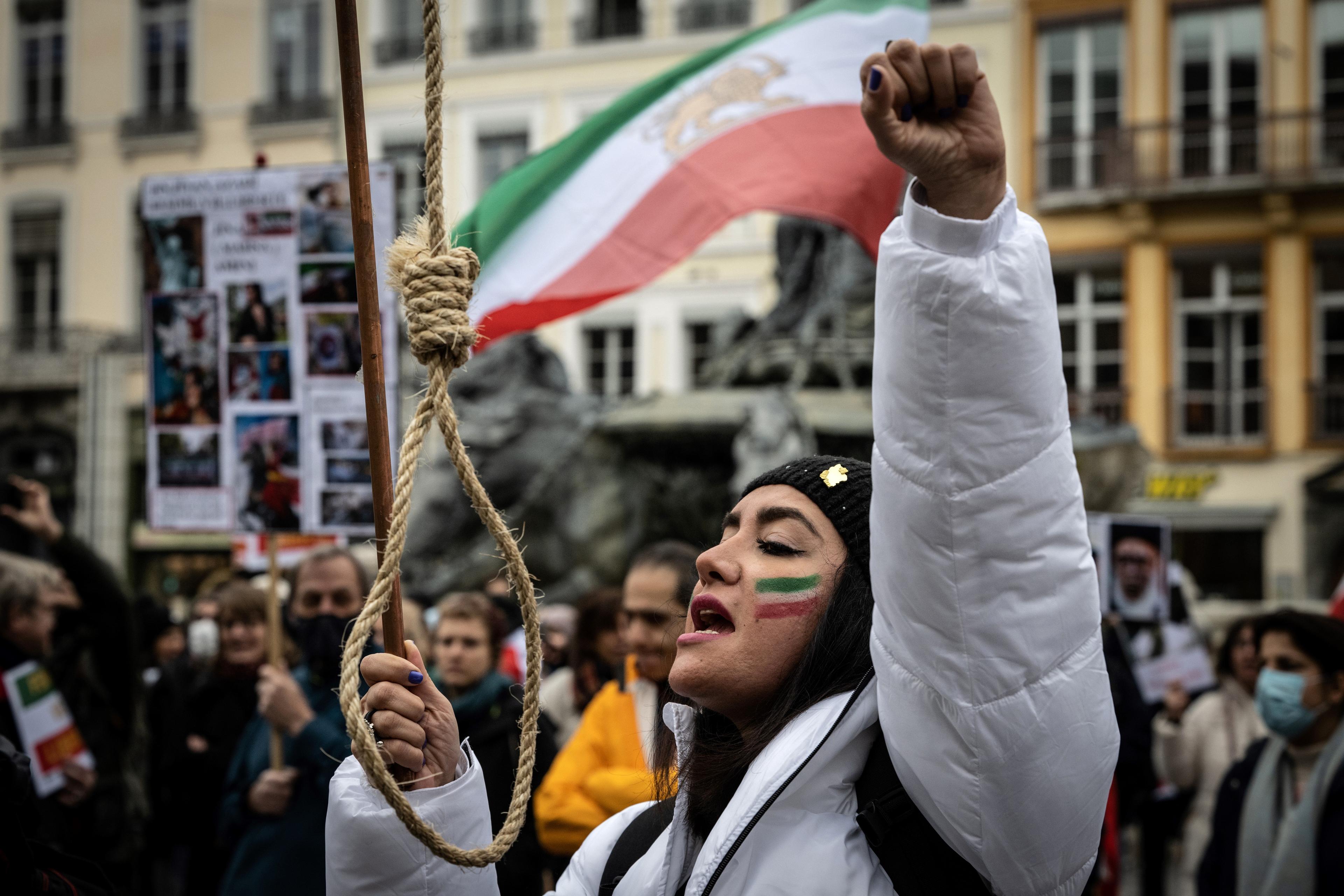 Protestująca kobieta podczas wiecu w jednej ręce trzyma sznur szubienicy. Drugie ramię unosi do góry. W tle flaga Iranu