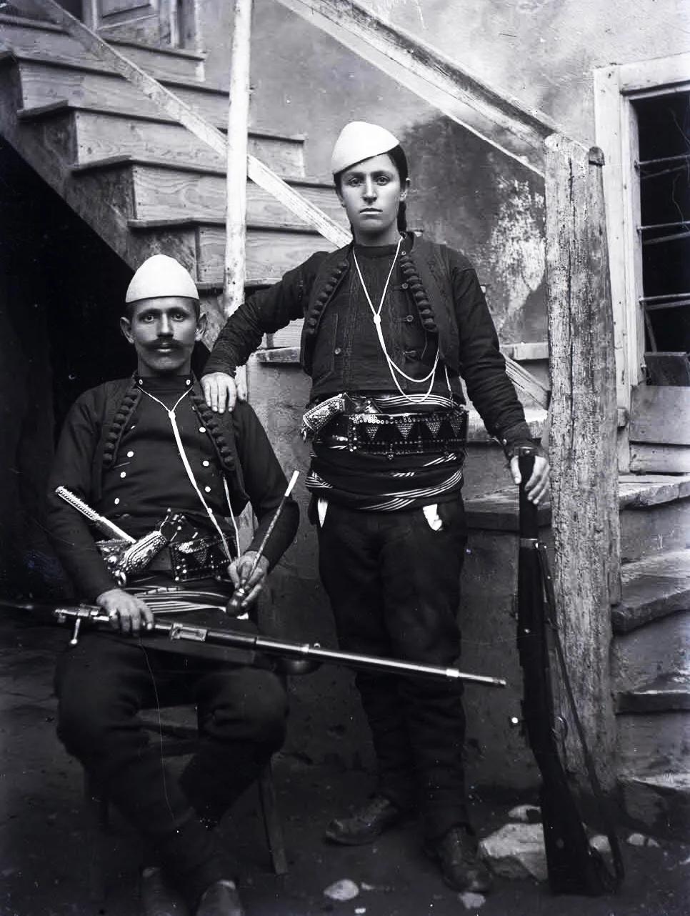 mężczyzna z wąsami i kobieta z warkoczem w tradycyjnych albańskich nakryciach głowy przypominających skorupkę jajka i w mundurach, pod bronią