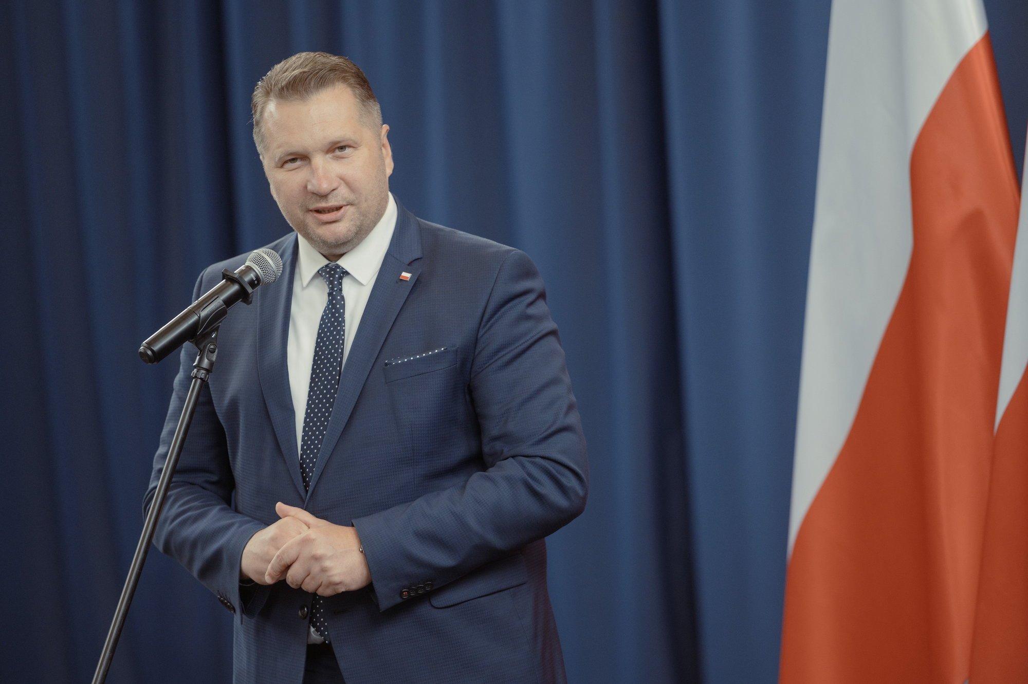 Minister Edukacji Przemysław Czarnek przemawia do mikrofonu ubrany w szaroniebieski garnitur i granatowy krawat w białe kropki.