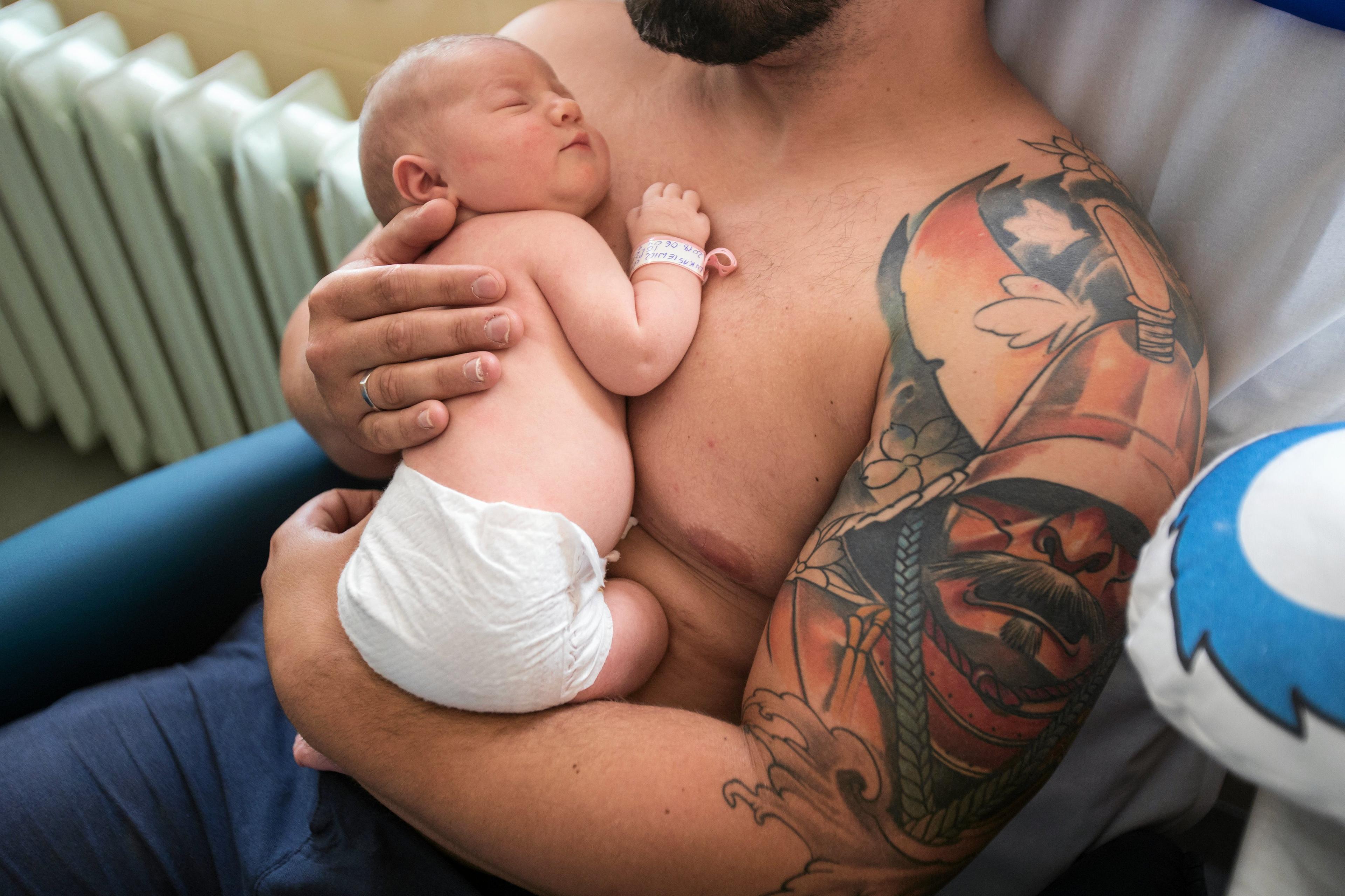 Mężczyzna z nagim torsem przytula noworodka. Na lewym ramieniu ma tatuaż przedstawiający wojownika
