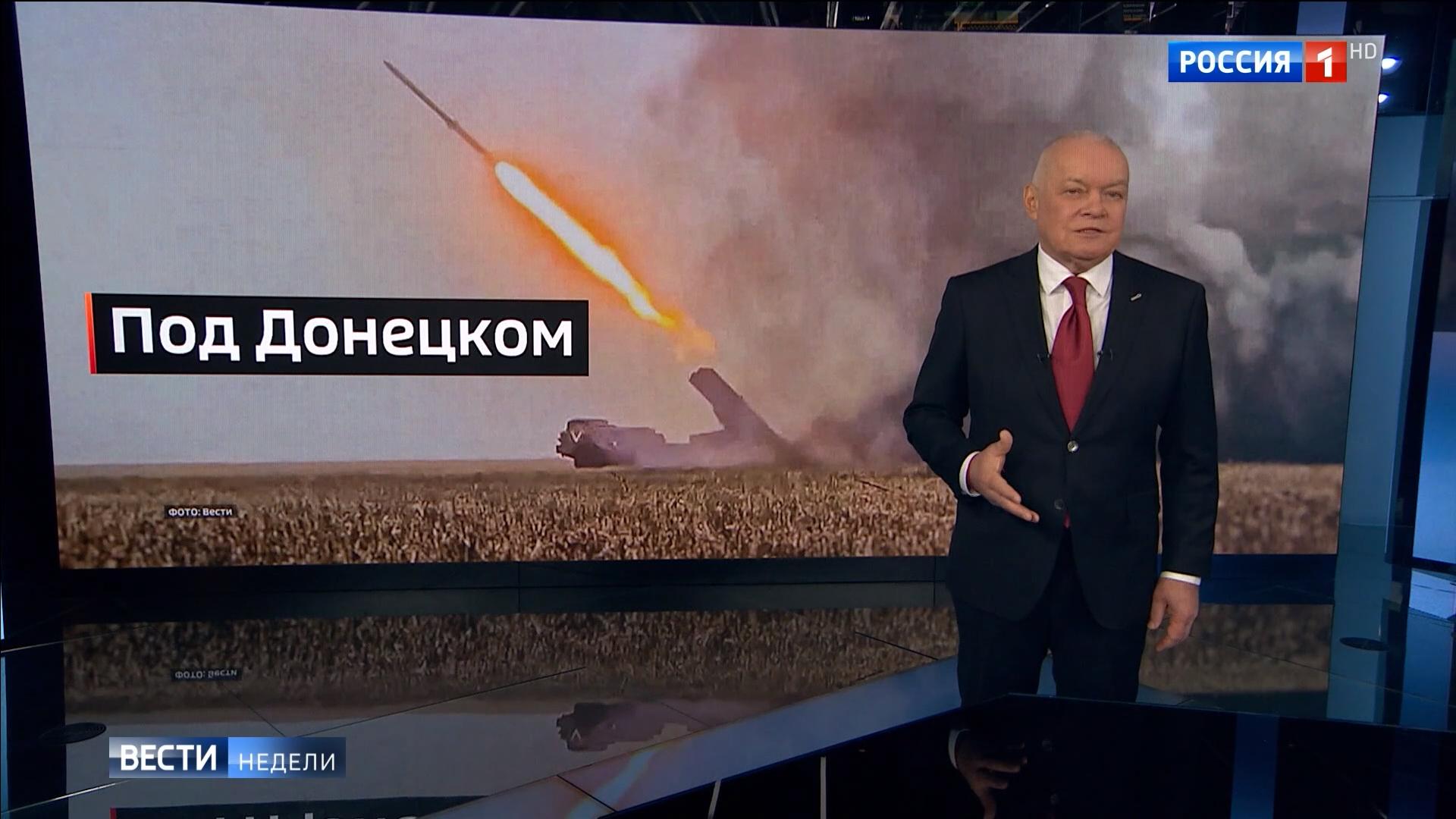 Starszy pan w telewizyjnym studio na tle zdjęcia ostrzału i rosyjskiego napisu 'pod Donieckiem"