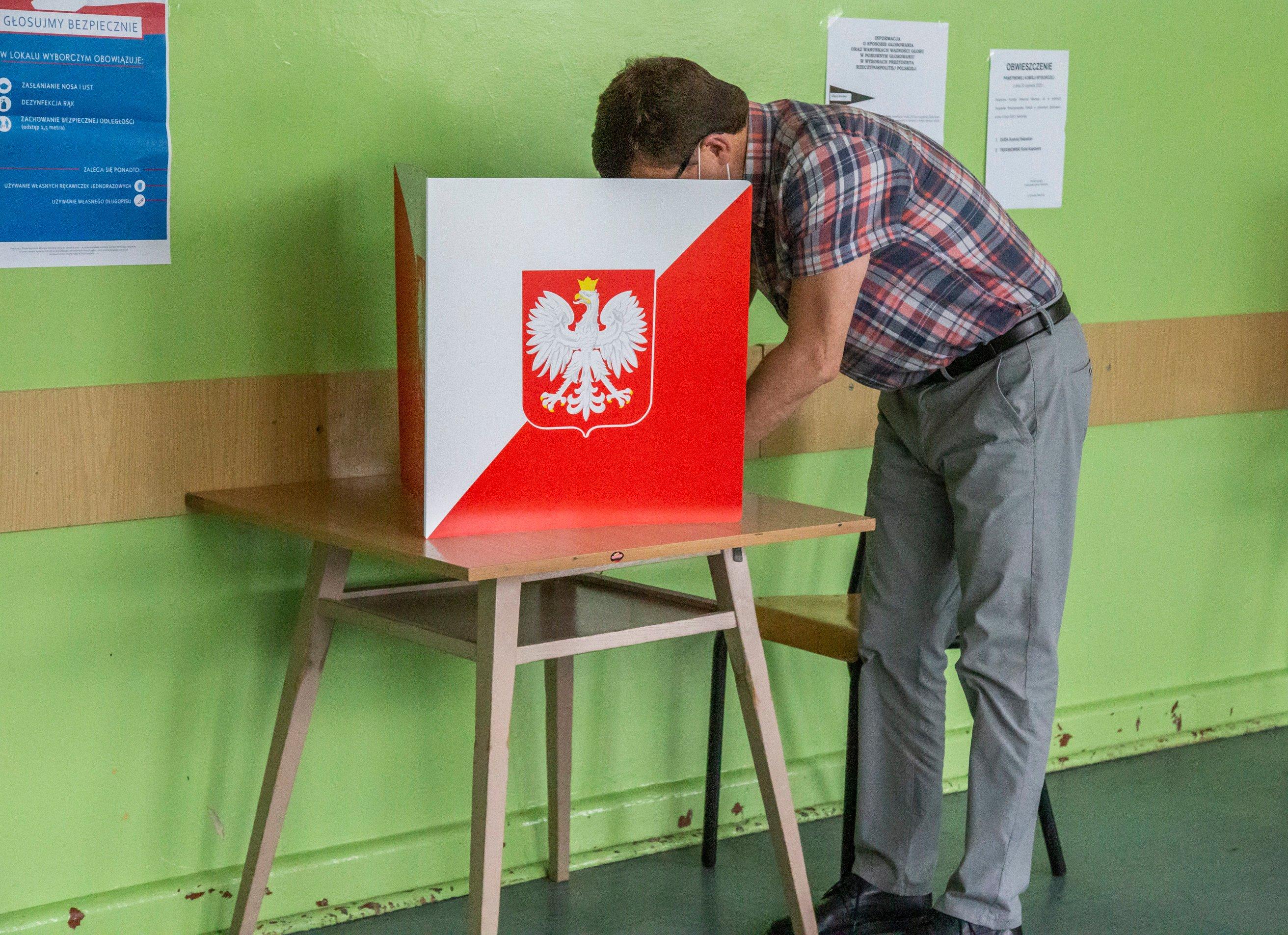 na zdjęciu mężczyzna pochyla się nad stolikiem osłoniętym kartonem z godłem Polski, żeby zaznaczyć głos na karcie wyborczej