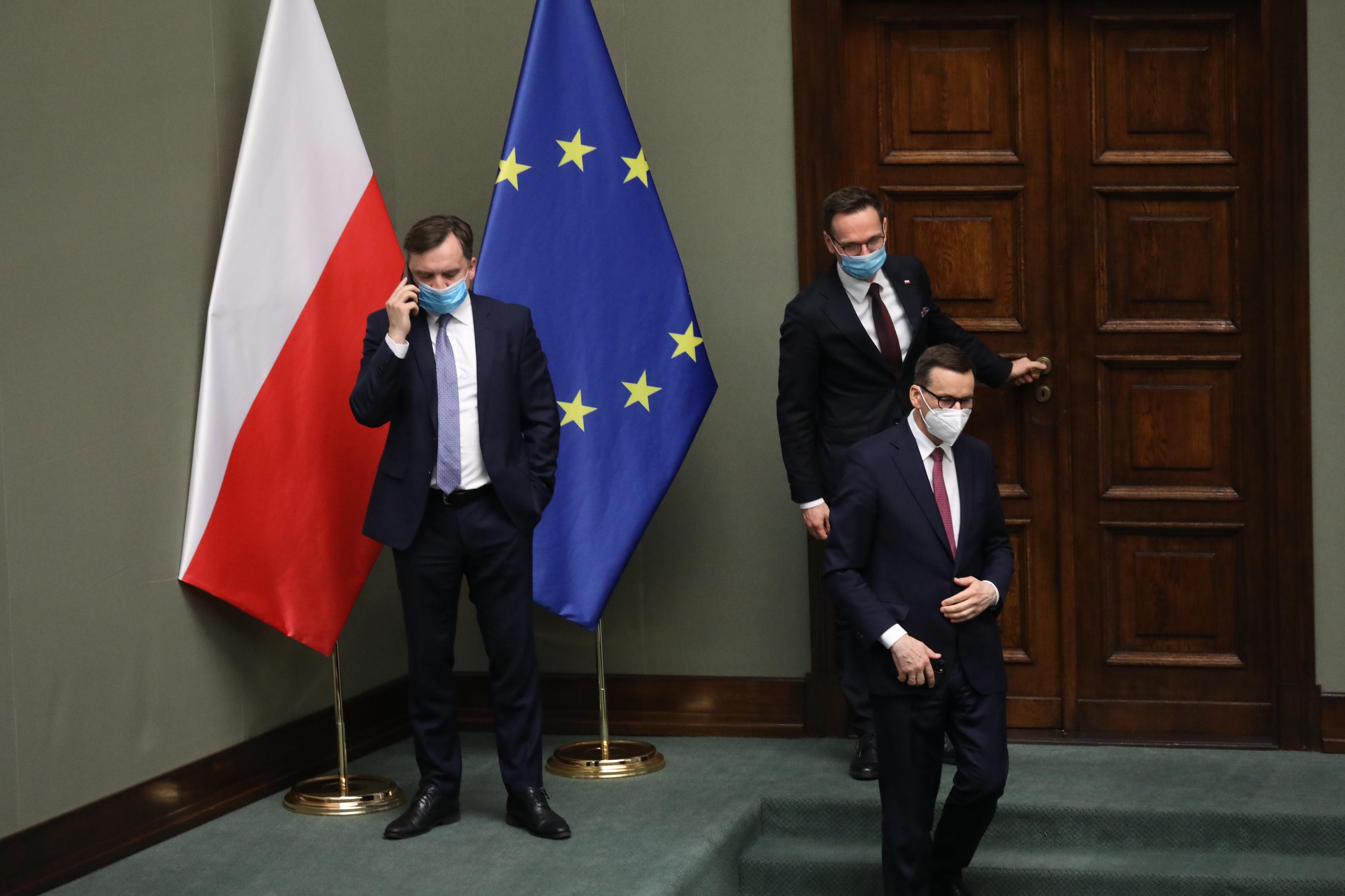 Zbigniew Ziobro w maseczce rozmawia przez telefon komórkowy w Sejmie, stojąc koło flagi Polski i Unii Europelskiej. Obok przechodzi premier Morawiecki, też w maseczce