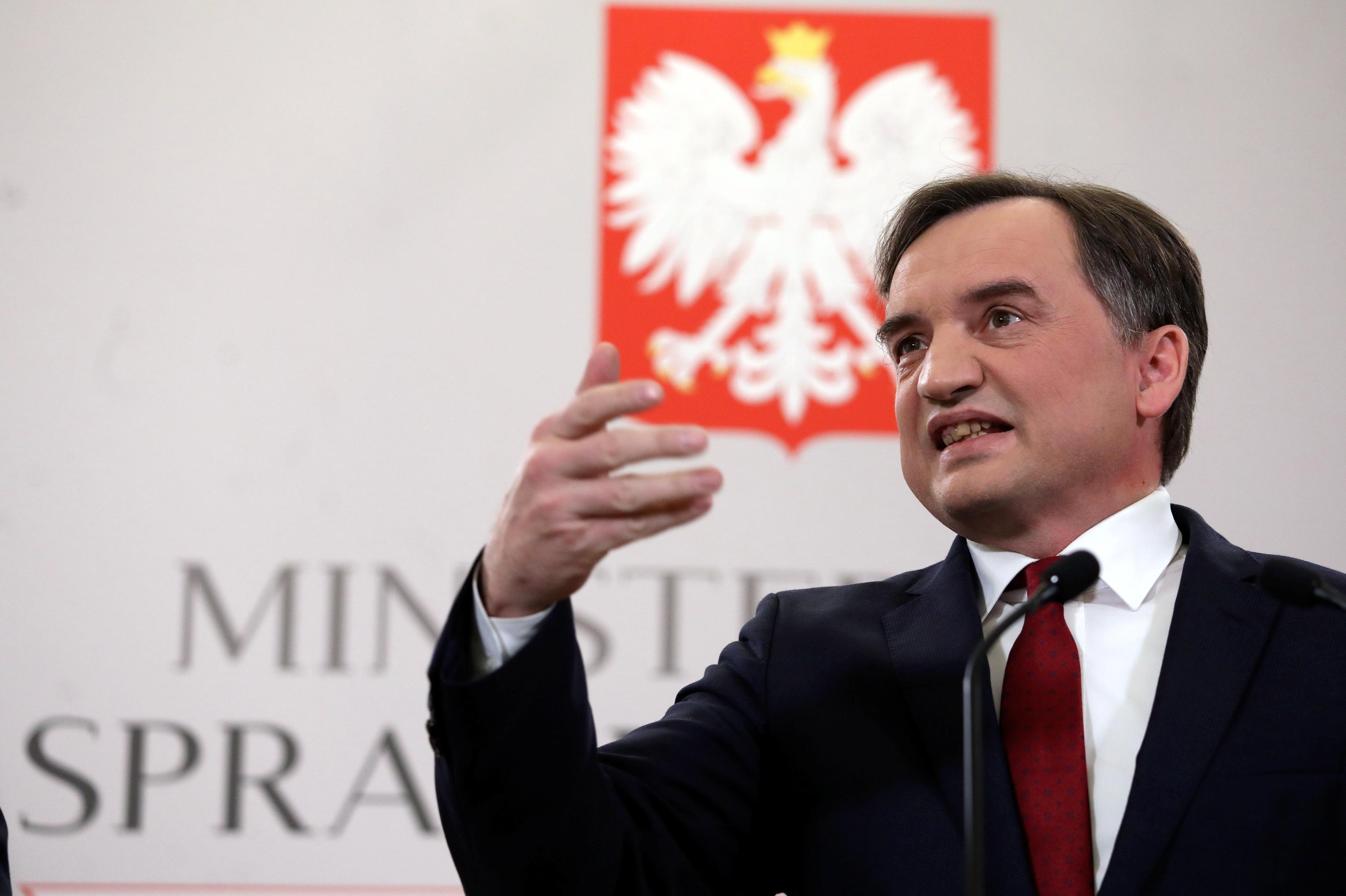 Minister Sprawiedliwości przemawia i ekspresyjnie gestykuluje prawą ręką na tle godła Polski