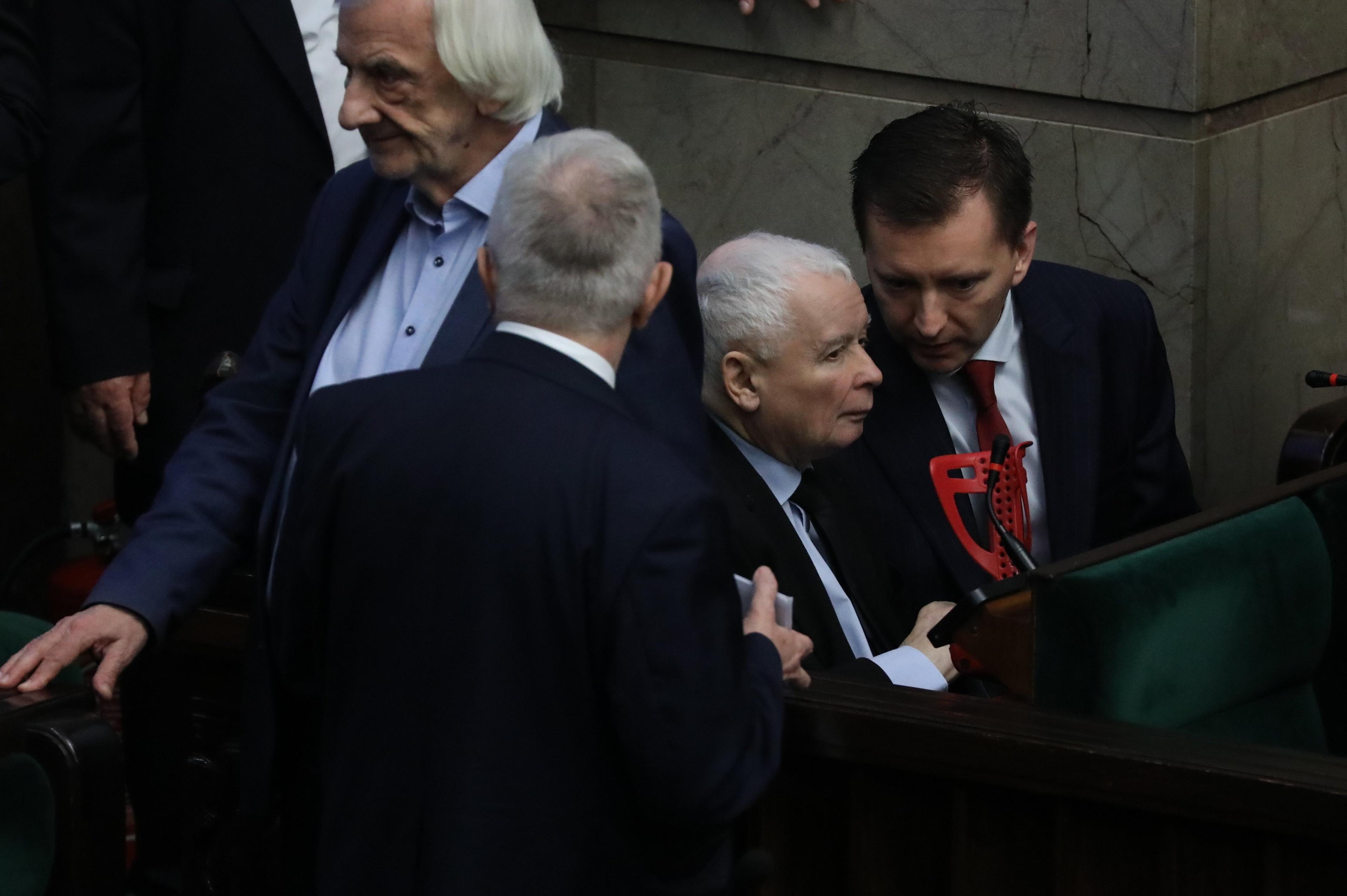 Jarosław Kaczyński otoczony przez posłów w ławach sejmowych. Przed nim oparte o tył fotela kule