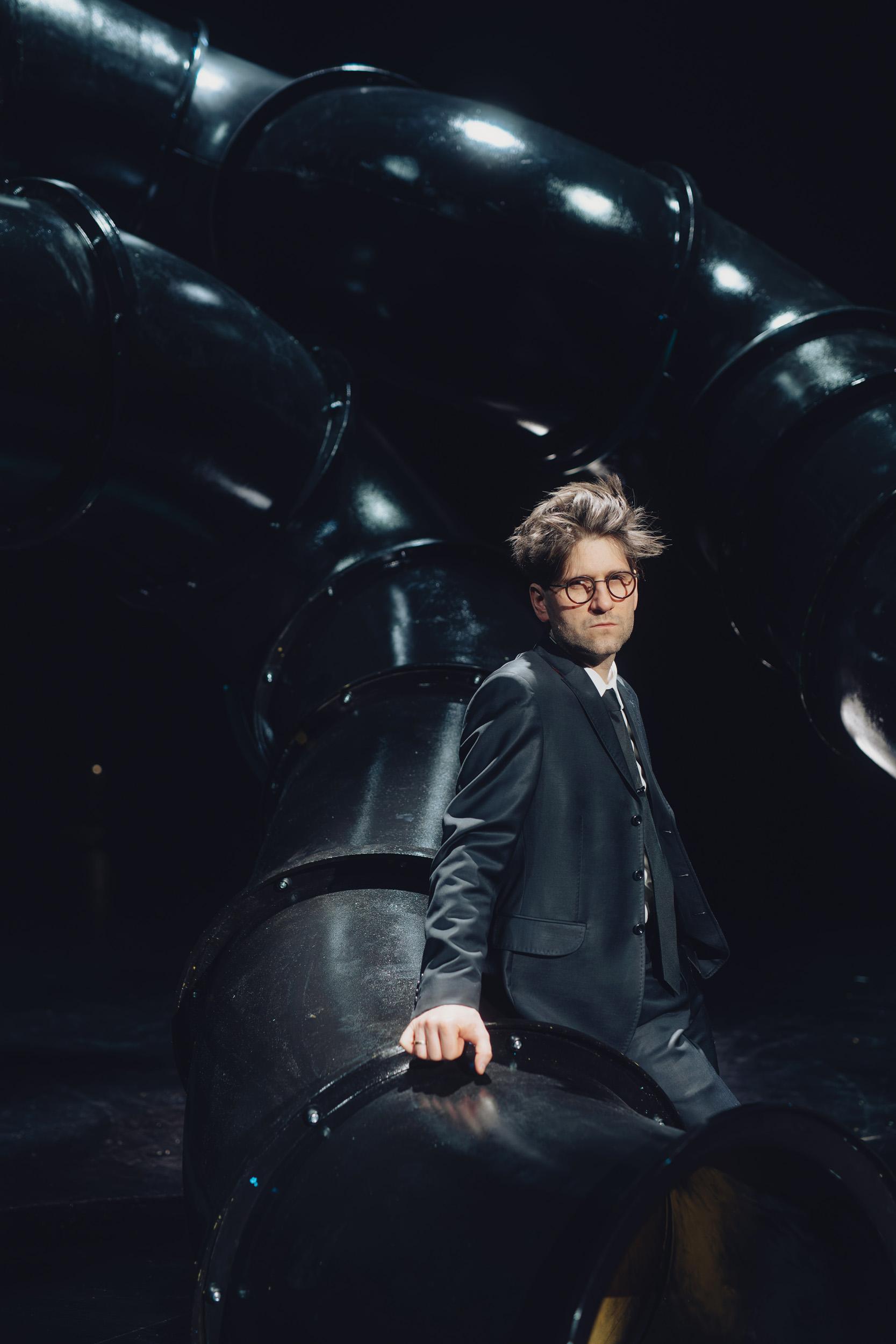 portret Mateusza Pakuły na tle scenografii teatralnej, przedstawiającej wielkie metalowe czarne rury