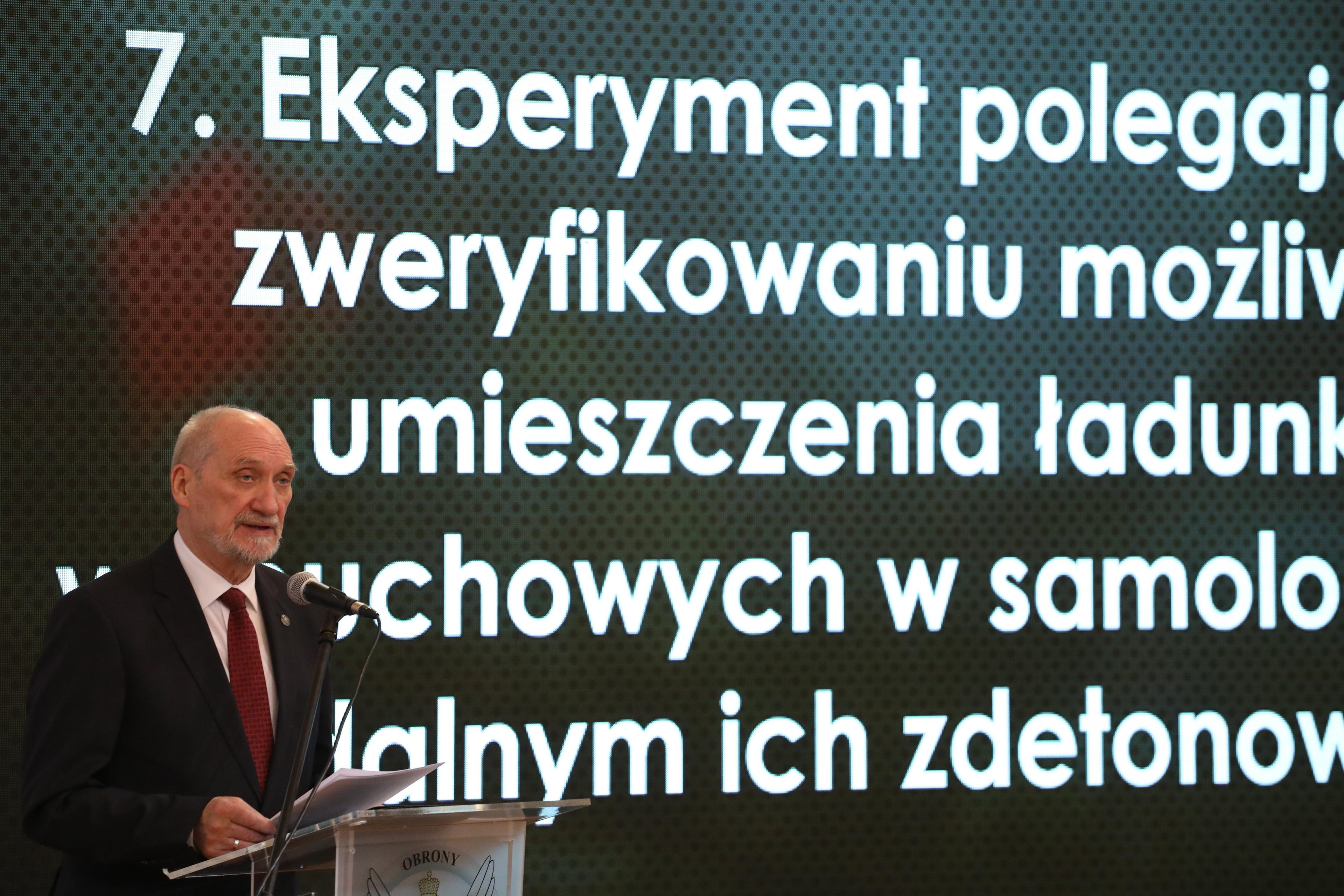 Mężczyzna, Antoni Macierewicz, występuje na scenie na tle ekranu z wyświetlonym raportem