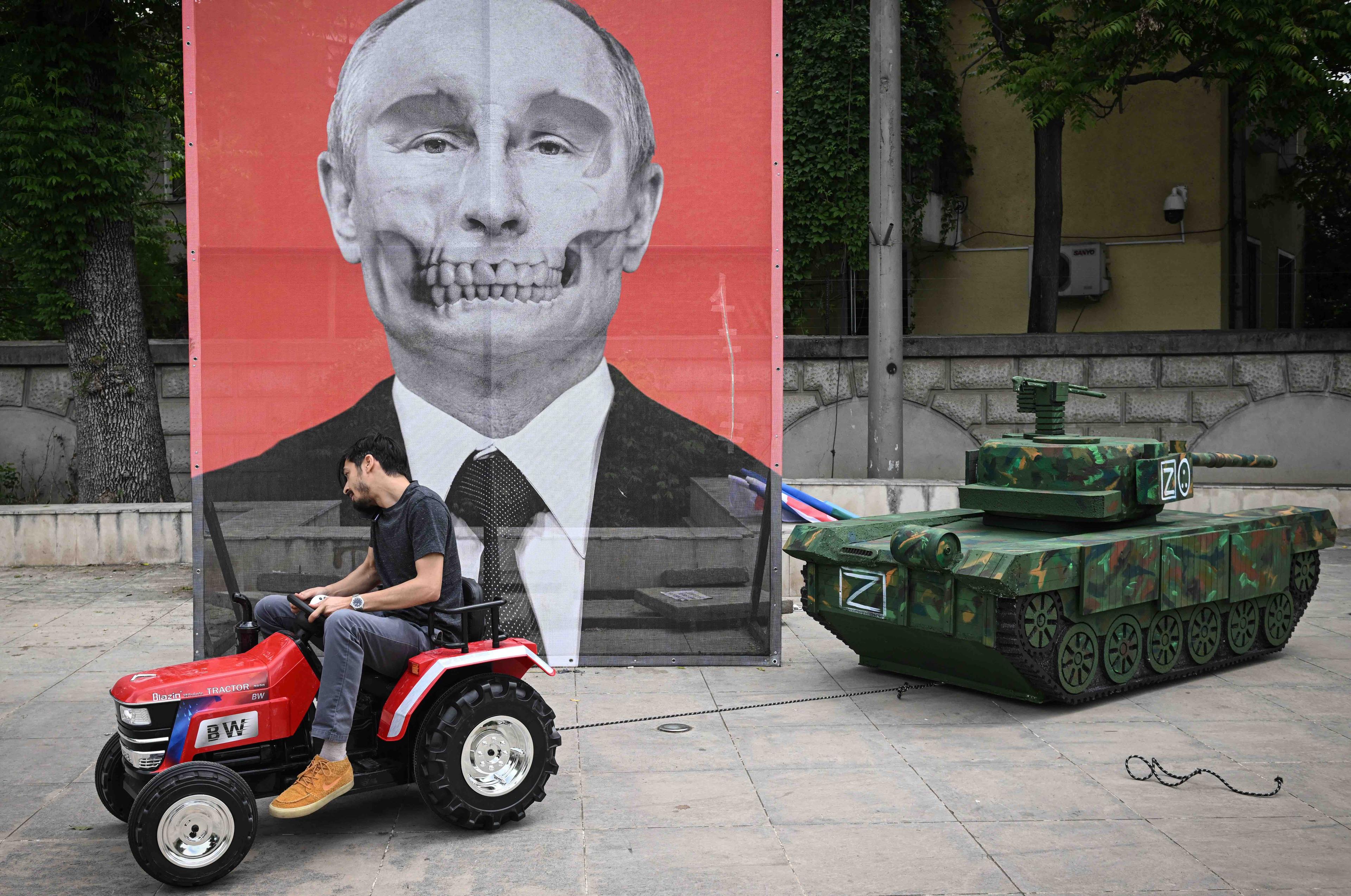 Mężczyzna prowadzi ogrodowy traktor, który ciągnie makietę rosyjskiego czołgu. W tle widać wielki portret Putina, którego twarz przypomina trupią czaszkę.