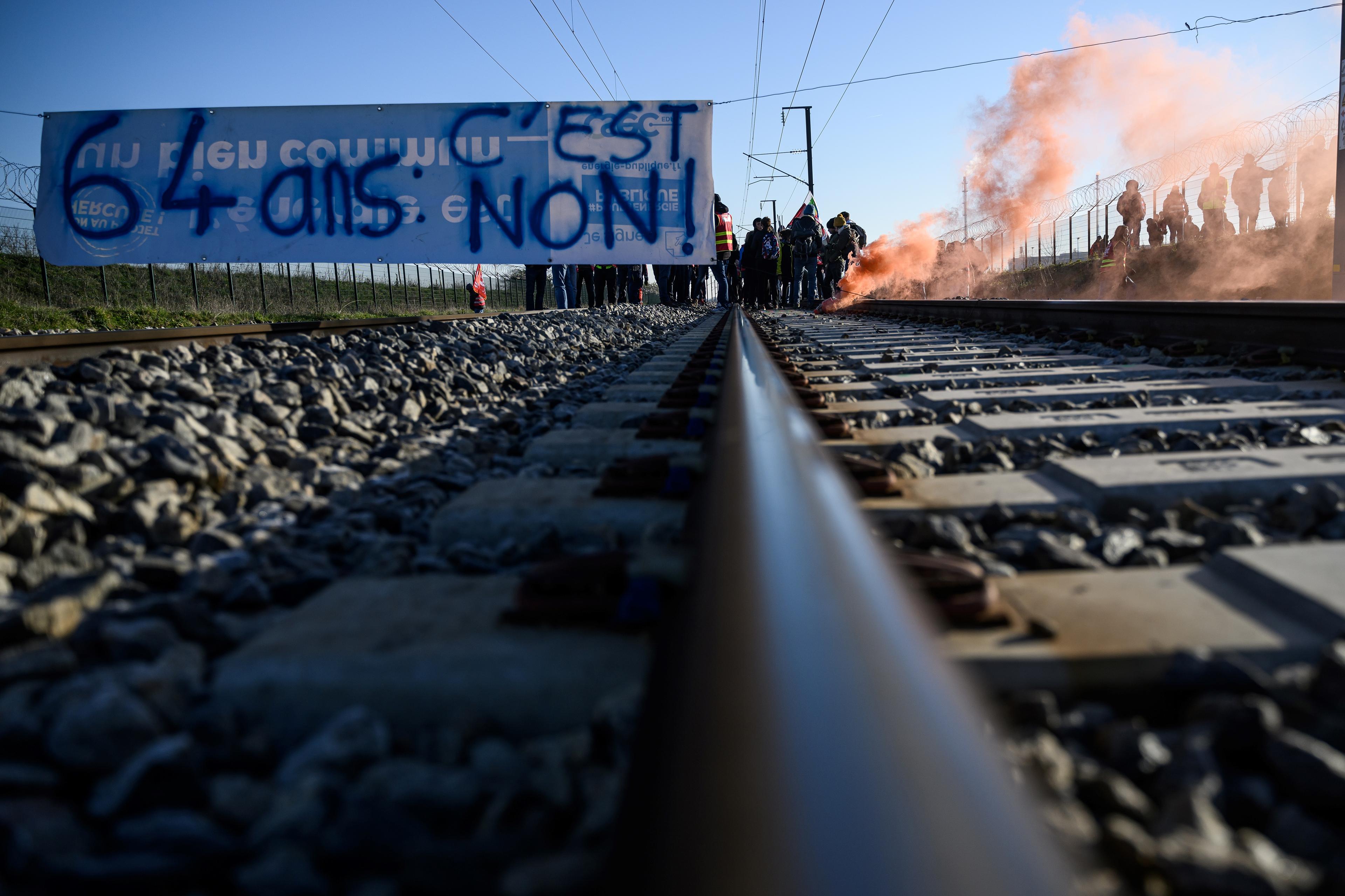 Protesters stand on the rail tracks behind a banner reading "64 years old, it's no" during an action called by the French union General Confederation of Labour (CGT) to protest against a deeply unpopular pensions overhaul in Donges, Western France on February 8, 2023. - France braced on February 7, 2023 for new strikes and mass demonstrations against French President's proposal to reform French pensions, including hiking the retirement age from 62 to 64 and increasing the number of years people must make contributions for a full pension. (Photo by LOIC VENANCE / AFP)