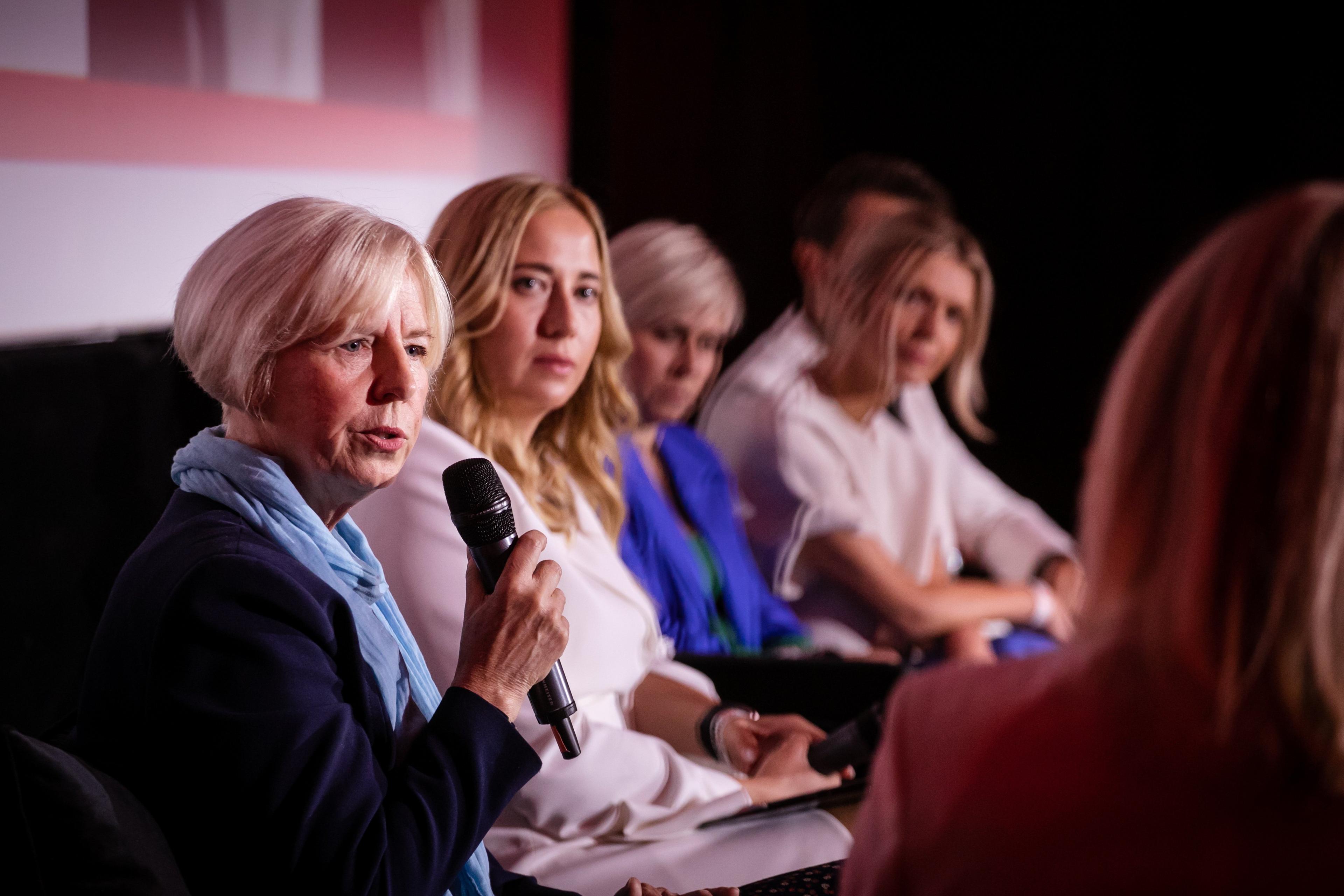 Blondynka w wieku średnim z mikrofonem w ręku siedząca w rzędzie wśród innych kobiet