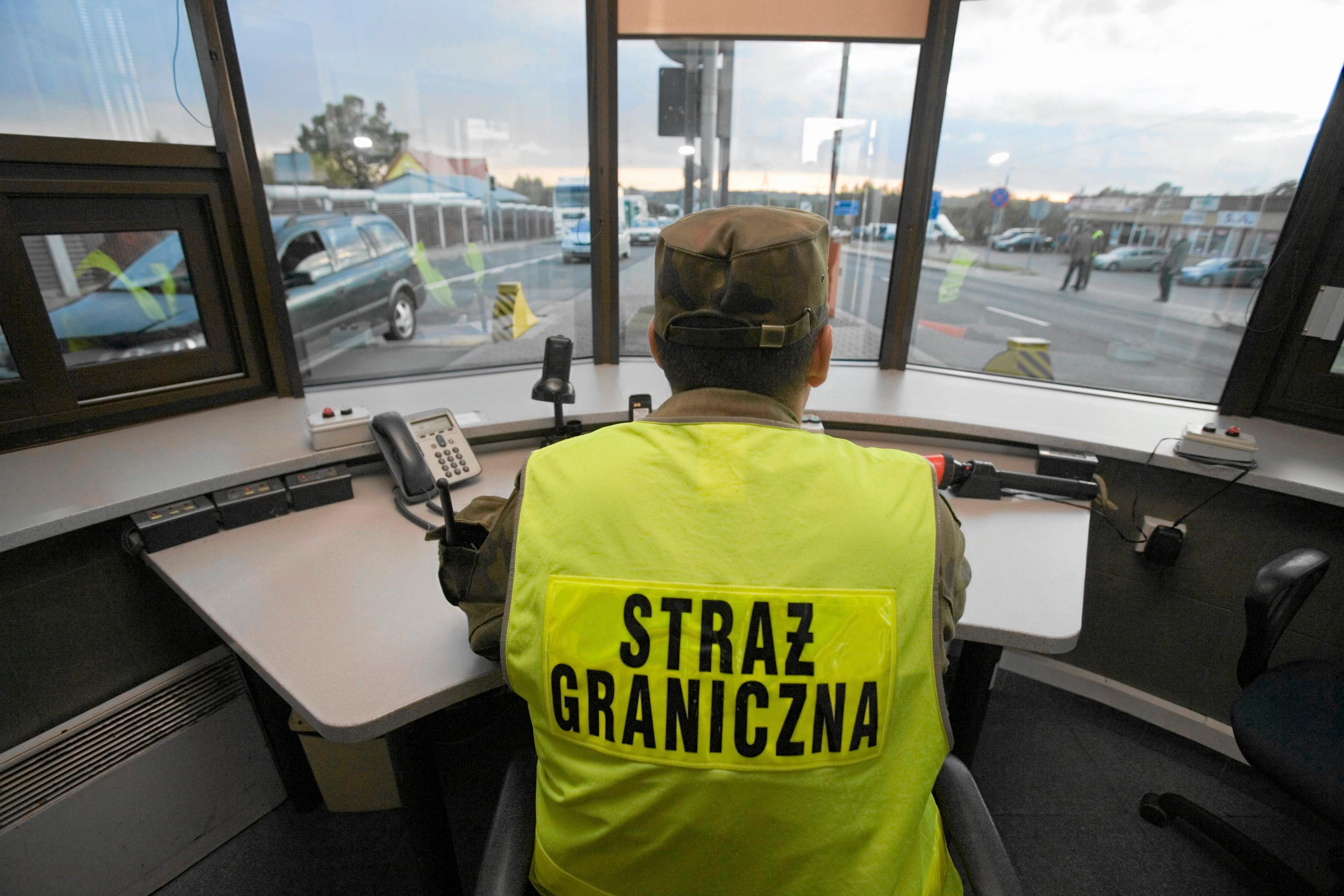 Na zdjęciu strażnik graniczny na przejściu w Kuźnicy Białostockiej. Mężczyzna w żółtej kamizelce i mundurze siedzi tyłem do obiektywu i kieruje ruchem drogowym na przejściu granicznym.