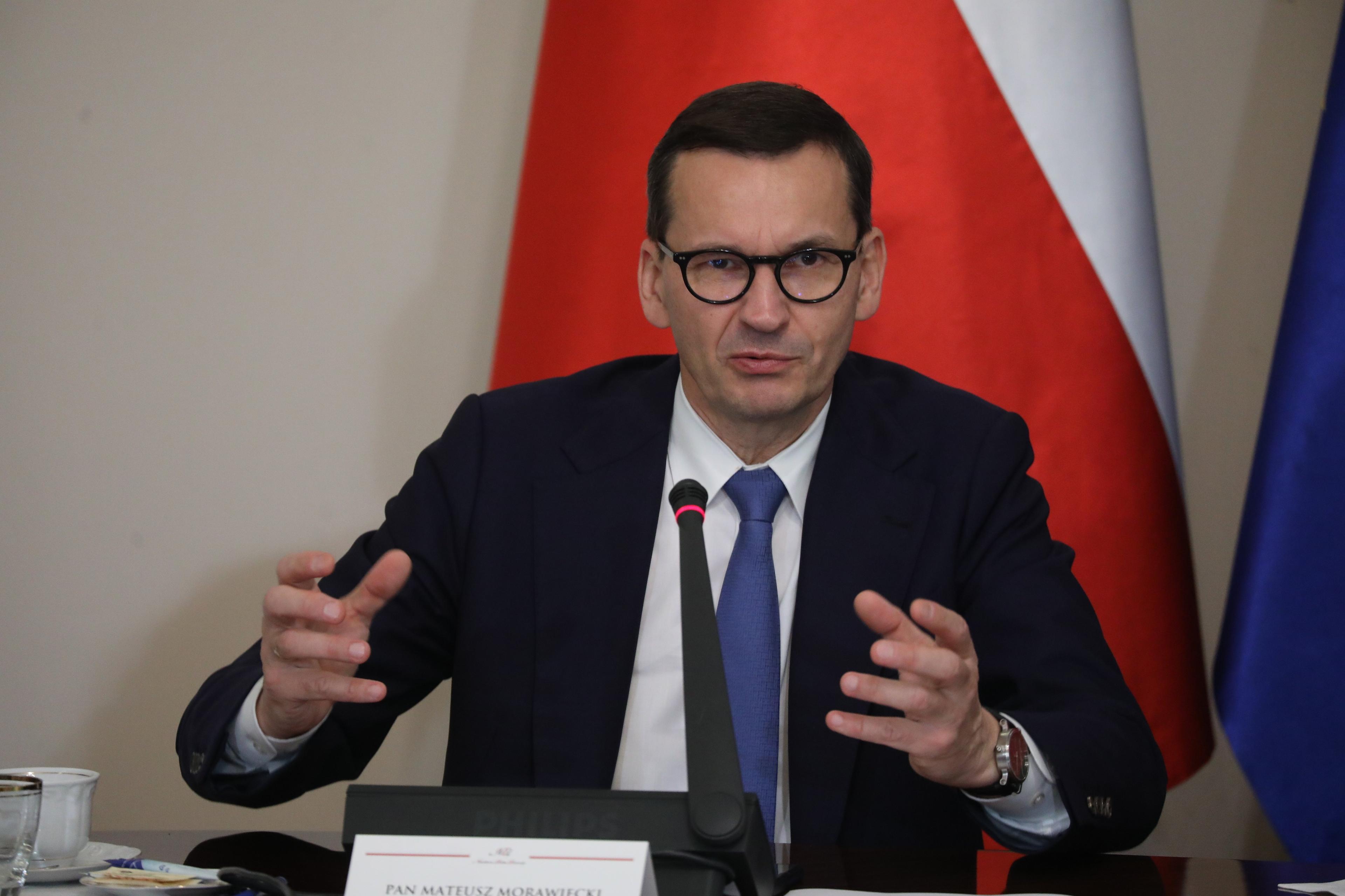 Premier Mateusz Morawiecki na tle polskiej flagi mówi do mikrofonu i gestykuluje