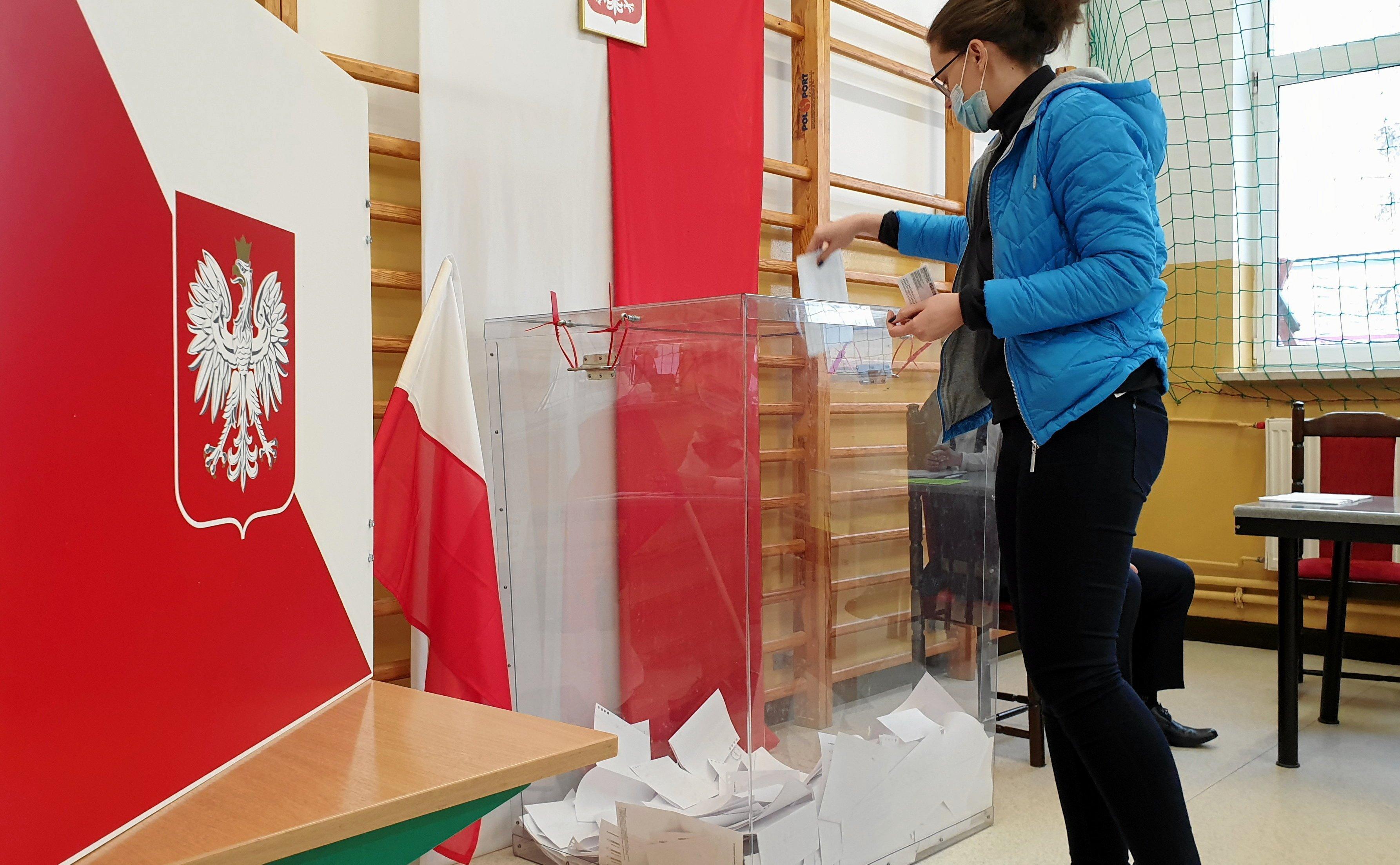 Kobieta wrzuca kartkę do przezroczystej urny wyborczej, w tle polska flaga