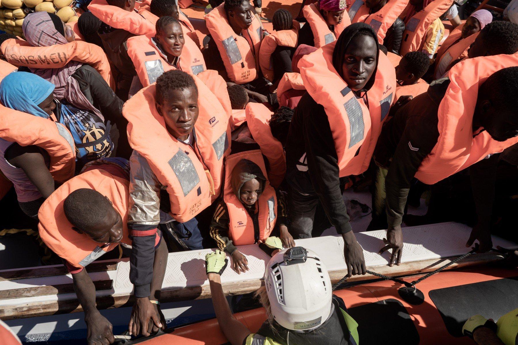 Grupa osób, kobiet i mężczyzn, w pomarańczowych kapokach wchodzą na pokład statku ratowniczego