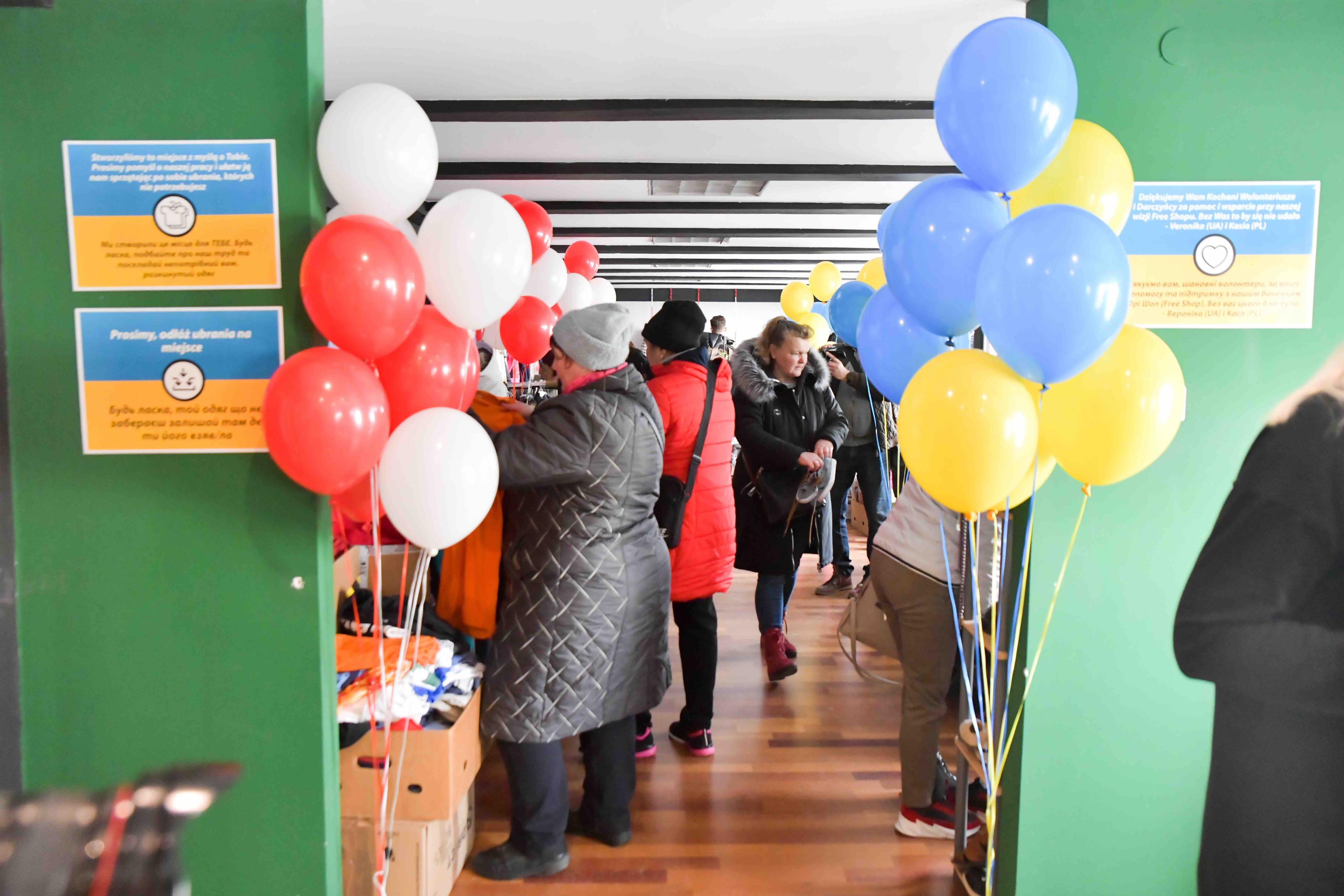 Wejcie do sklepu z bezpłatną pomocą dla uchodźców z Ukrainy, po obu stronach weśjcia baloniki w kolorach flagi polskiej i ukraińskiej, w głębi osoby przeglądają rzeczy..