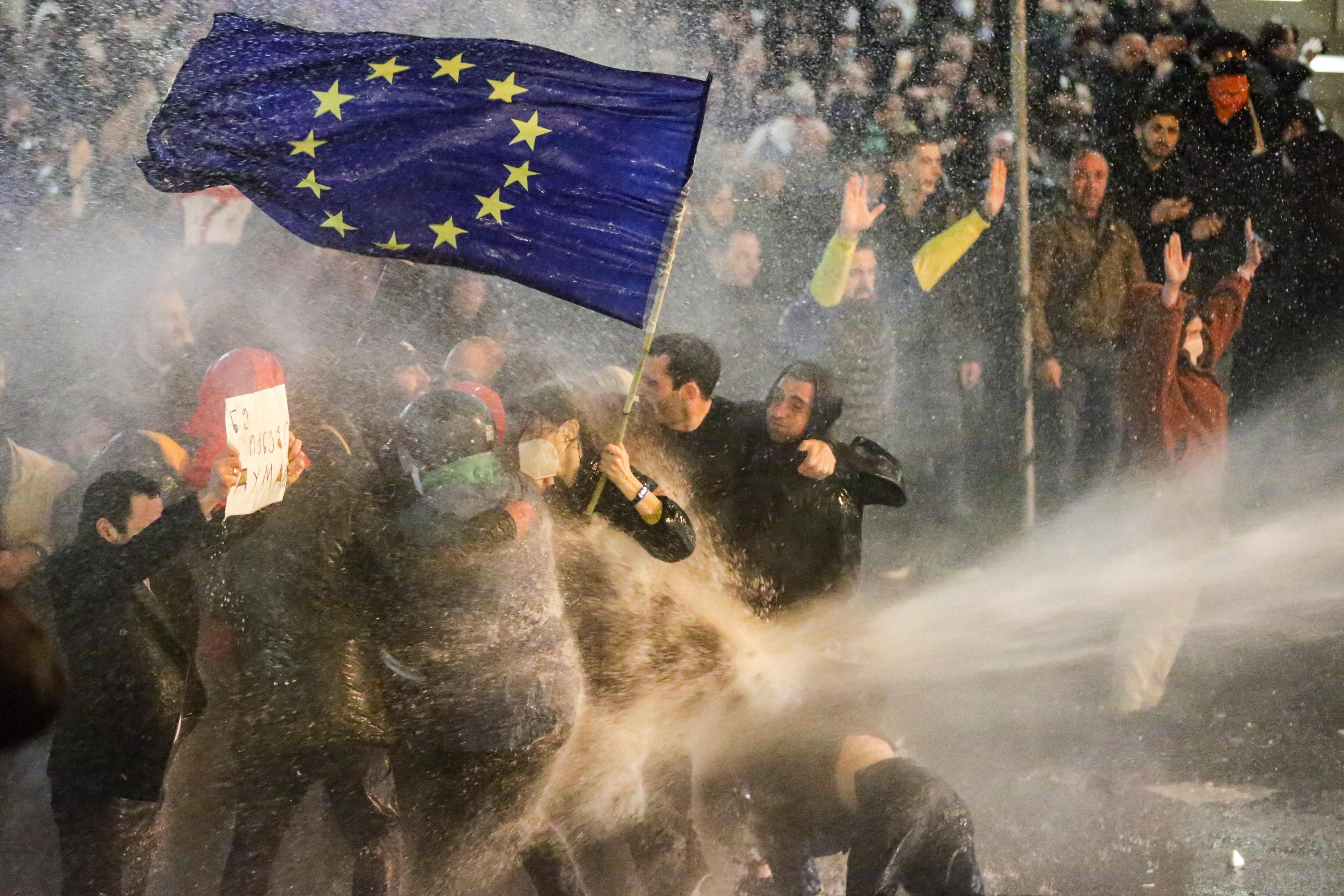 Protestujący niosący flagę Unii Europejskiej są opryskiwani przez armatki wodne podczas starć z policją