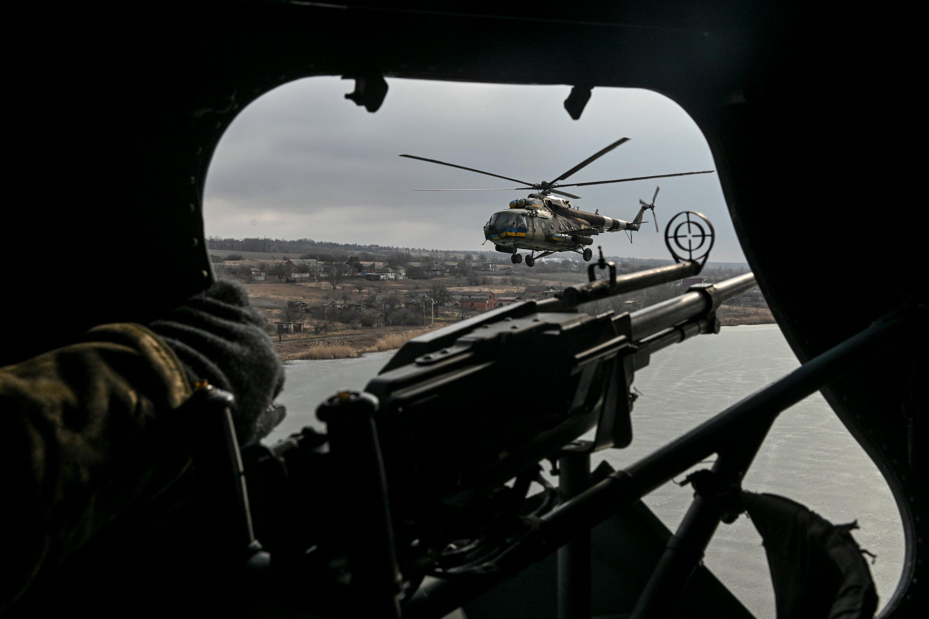 Zdjęcie wykonane z wnętrza helikoptera. Na pierwszym planie broń maszynowa wystawiona przez otwarty właz do helikoptera. Na dalszym planie inne helikopter lecący na zbiornikiem wodnym.
