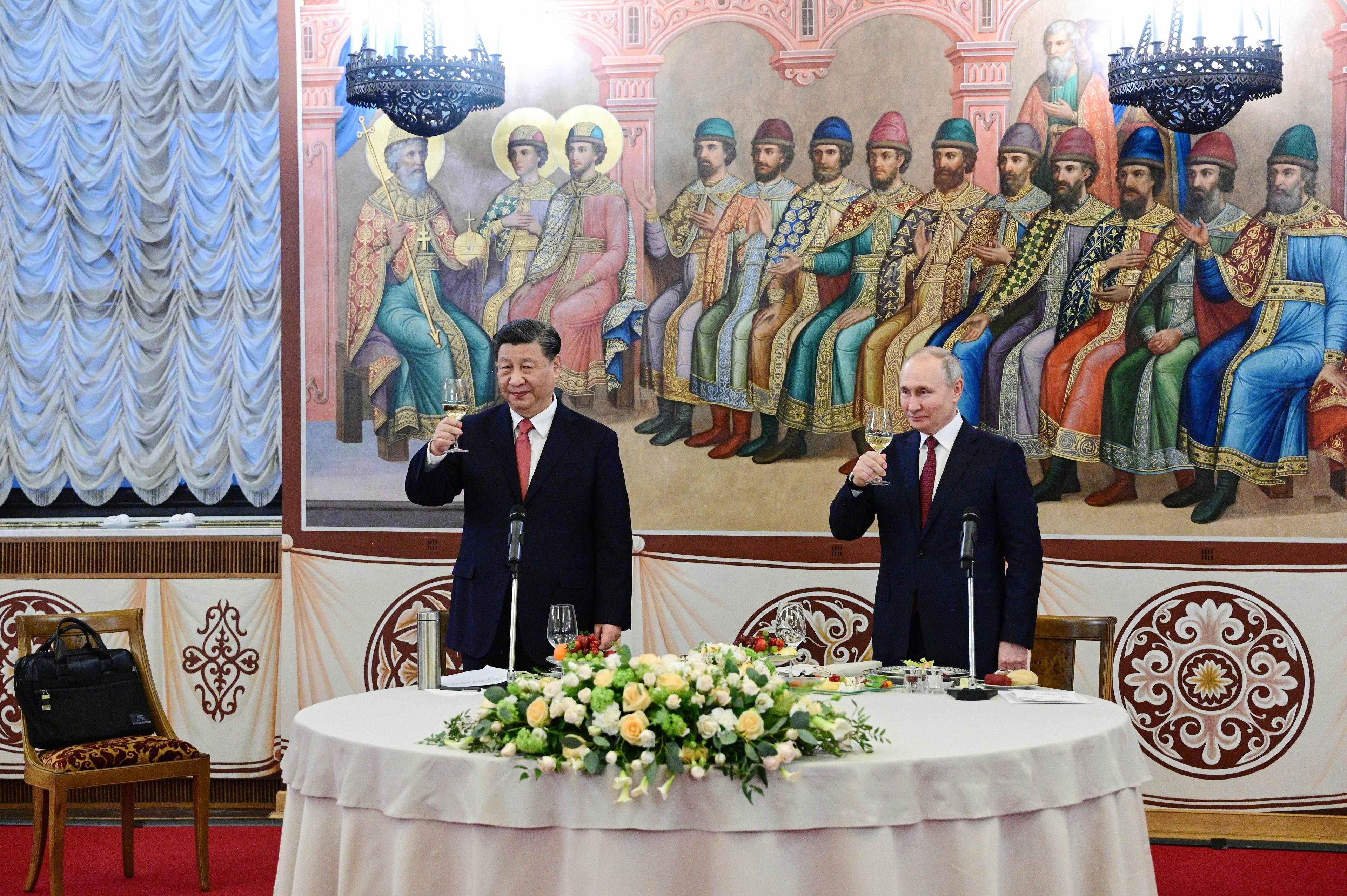 Władimir Putin i Xi Jinping stoją przy stole bankietowym z kieliszkami. Na stole żółte kwiaty, za politykami fresk