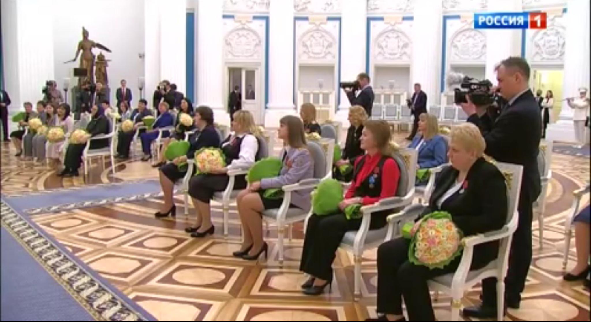 Kobiety z bukietami żółtych kwatów siedzą w ozdobnych fotelach rozstawionych w rzędach w sali pałacowej