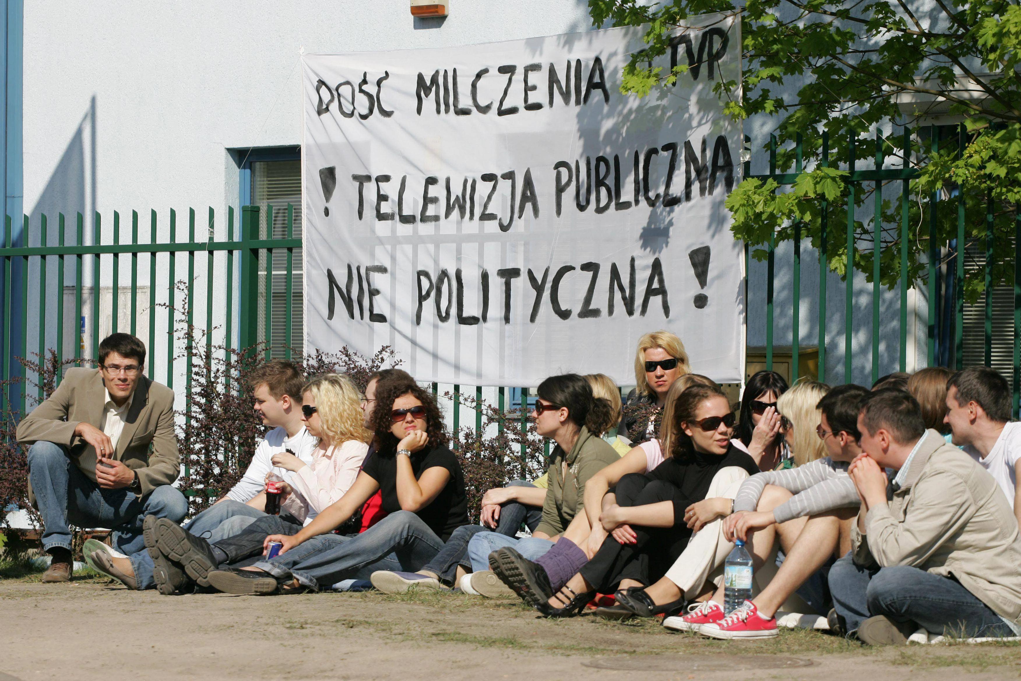 grupa ludzi siedząca przed transparentem domagającym się publicznej, nie politycznej telewizji