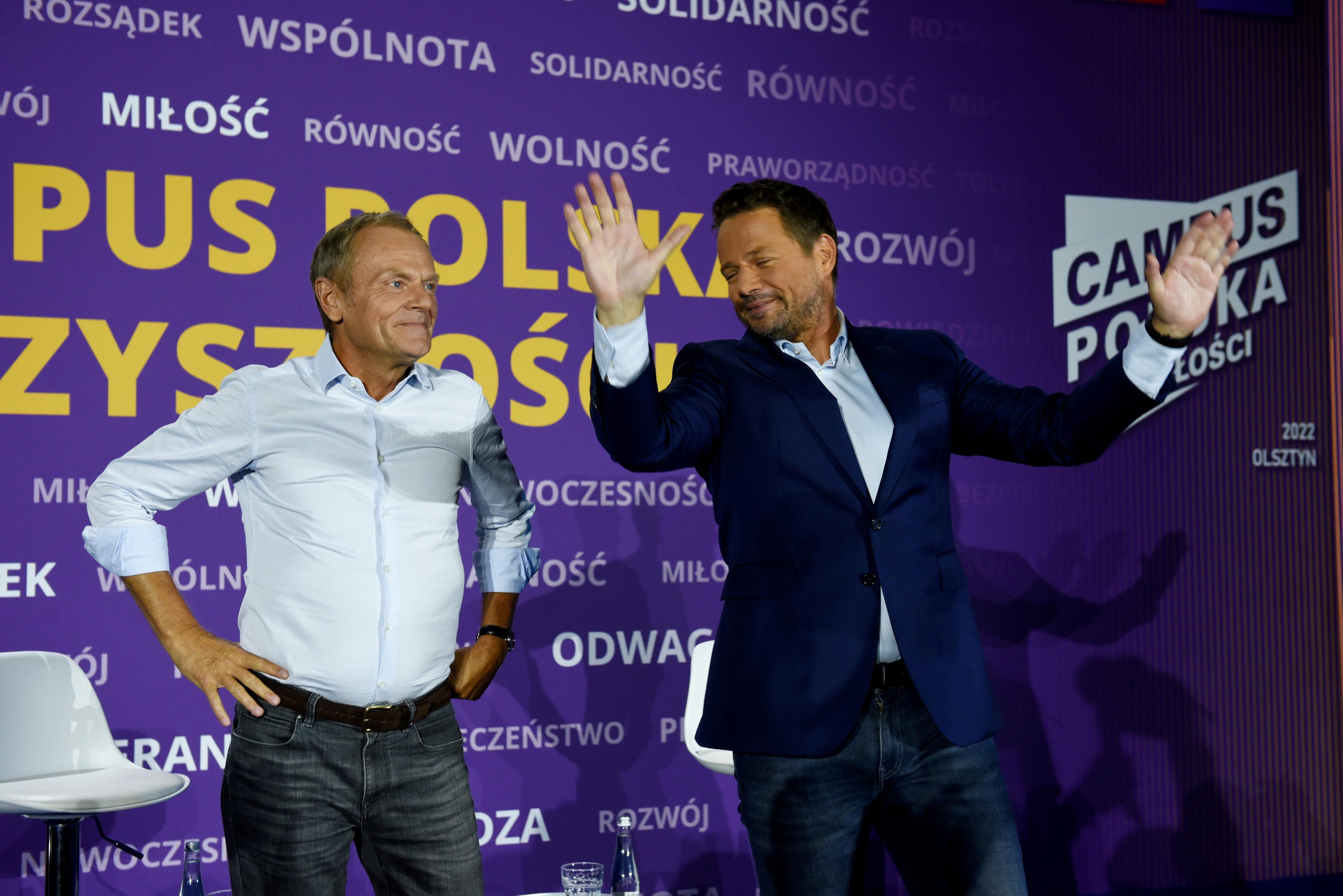 Donald Tusk w białej koszuli, i Rafał Trzaskowski w białej koszuli i ciemnej marynarce, stoją obok siebie, za nimi ścianka z logo Campusu Polska
