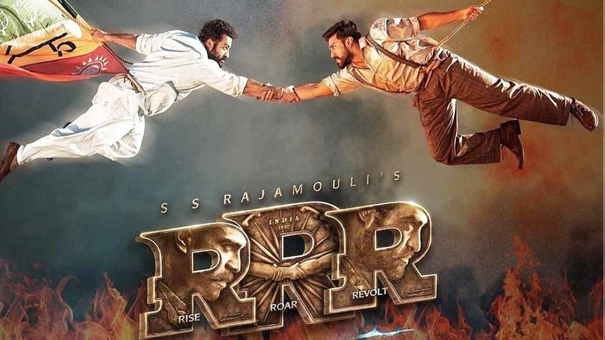 Plakat filmu „RRR”, przedstawiający dwóch bohaterów, zawieszonych na linach, którzy podają sobie w locie ręce ręce, jeden z nich trzyma indyjską flagę; pod spodem tytuł „RRR” i jego rozwinięcie w języku angielskim: rise, roar, revolt.