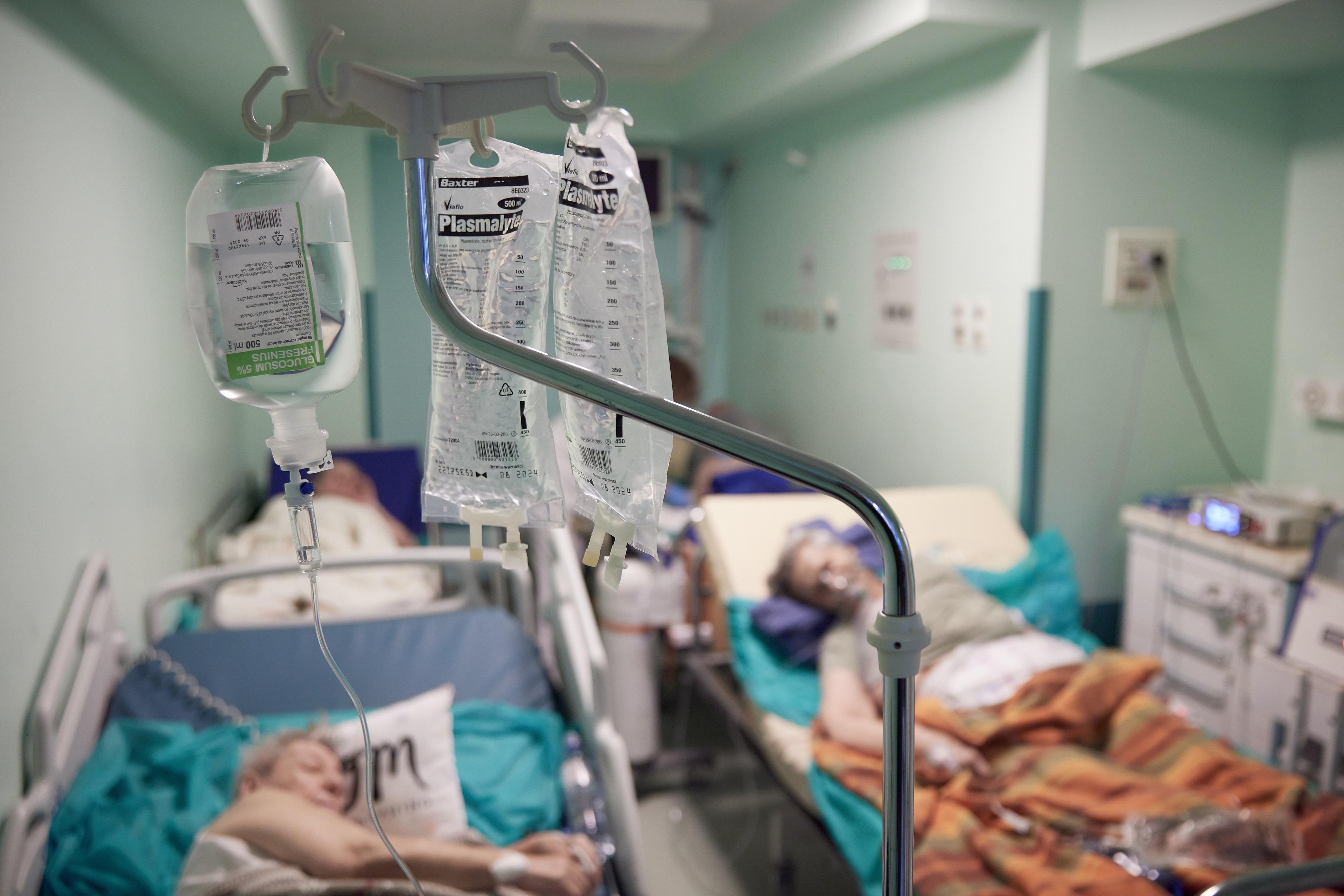 Na zdjęciu trzy łóżka szpitalne obok siebie, na nich lkudzie podłączeni do kroplówek