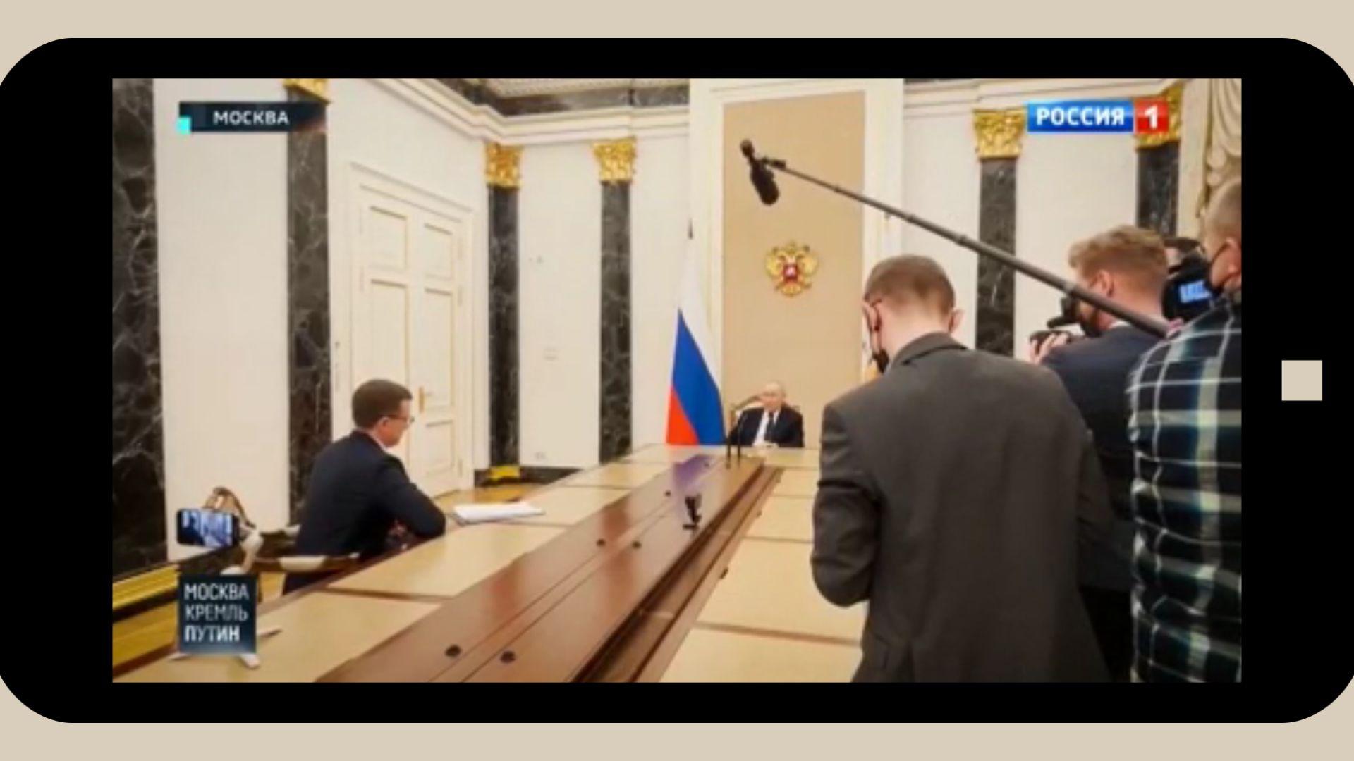 Grafika: w ramke telefinu komórkowego wklejony kadr z wywiadu Putina z Pawłem Zarubinem: Putin siedzi u szczytu bardzo długiego stołu. W połowie stołu - młody mężczyzna (Zarubin), na drugim końcu - kamerzyści i operatorzy dźwięku w maskach