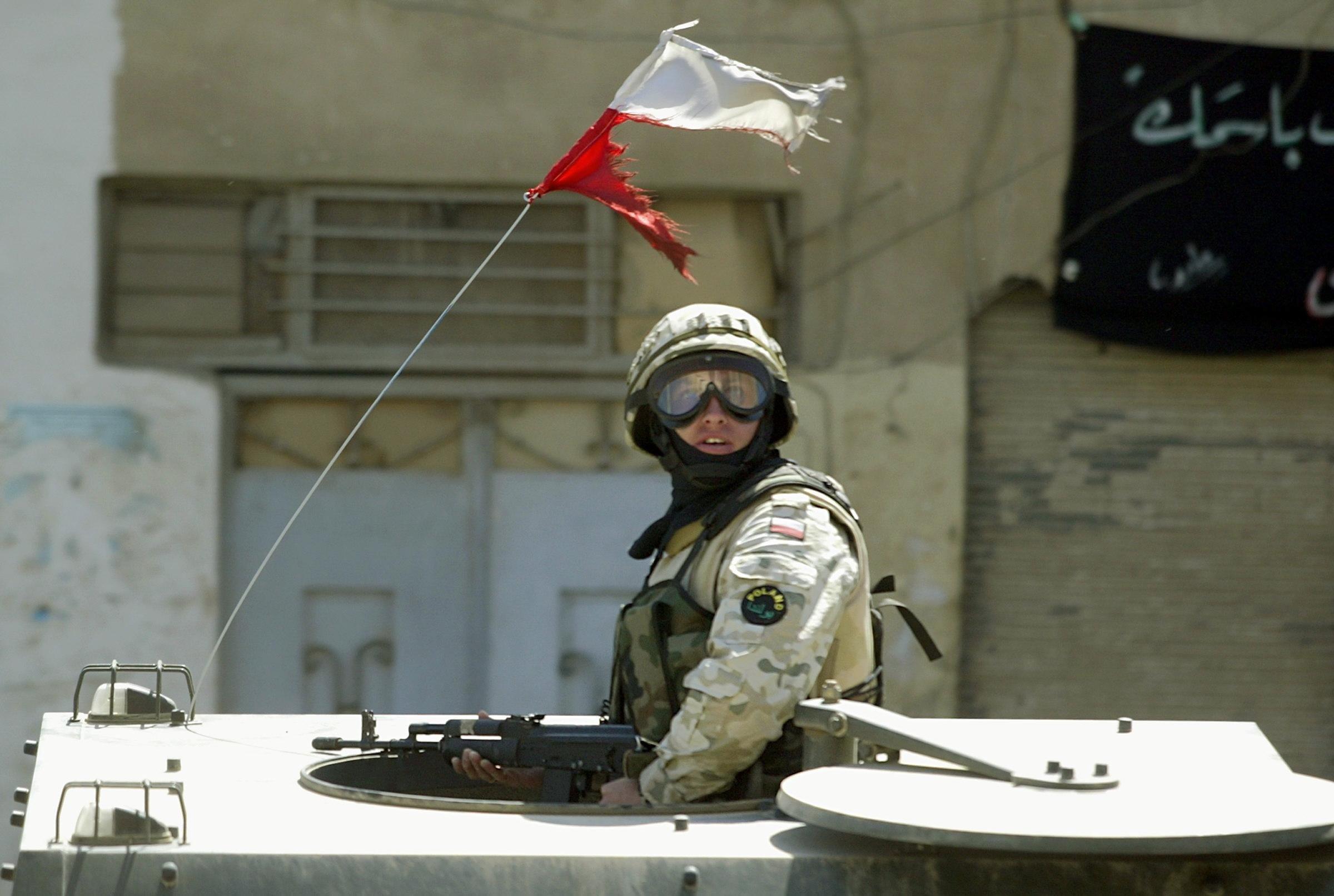 Polski żołnierz w hełmie i goglach w wieżyczce transportera opancerzonego na ulicy w irackim mieście. Nad głową żołnierza powiewa rozdarta polska flaga.
