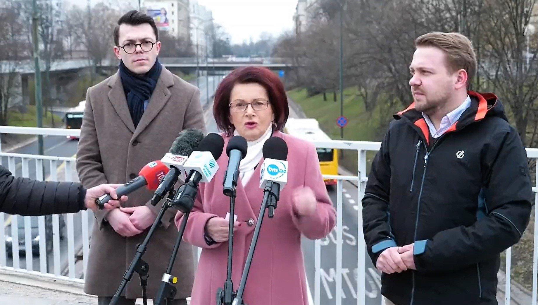 Kobieta na kładce nad Trasa Łazienkowską w Warszawie mówi do mikrofonów, po obu stronach dwaj mężczyźni