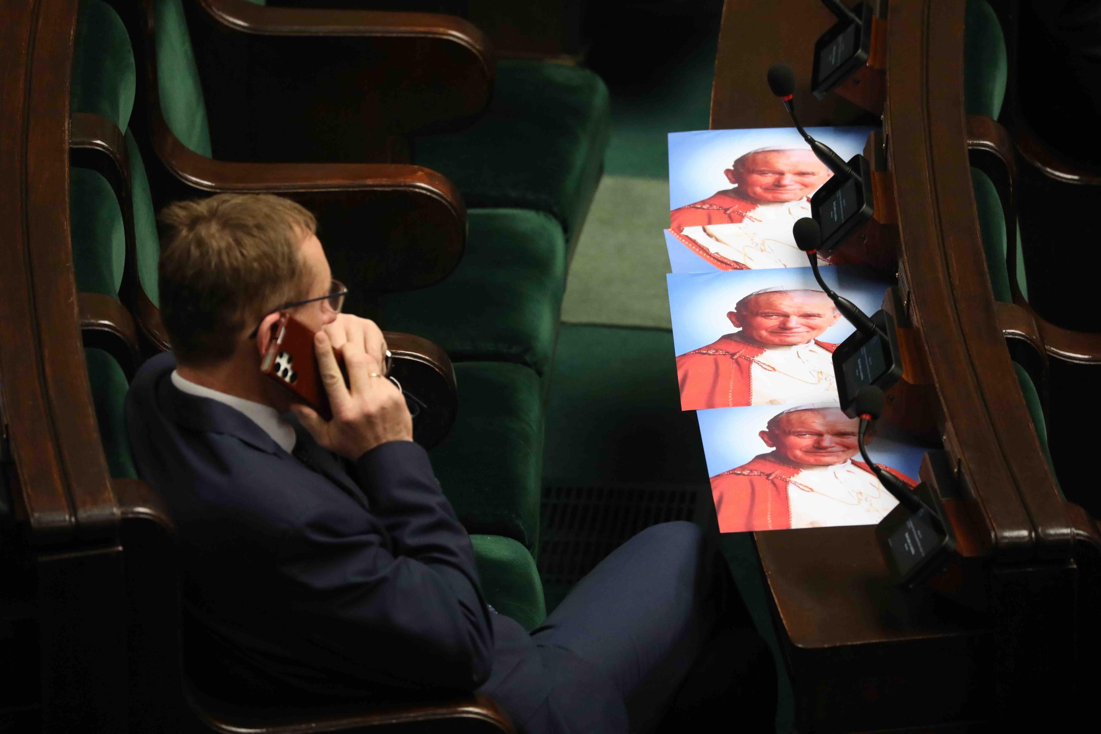 Portrety Jana Pawła II leżą w miejscach poselskich na sali Sejmu. Obok poseł rozmawia przez telefon.
