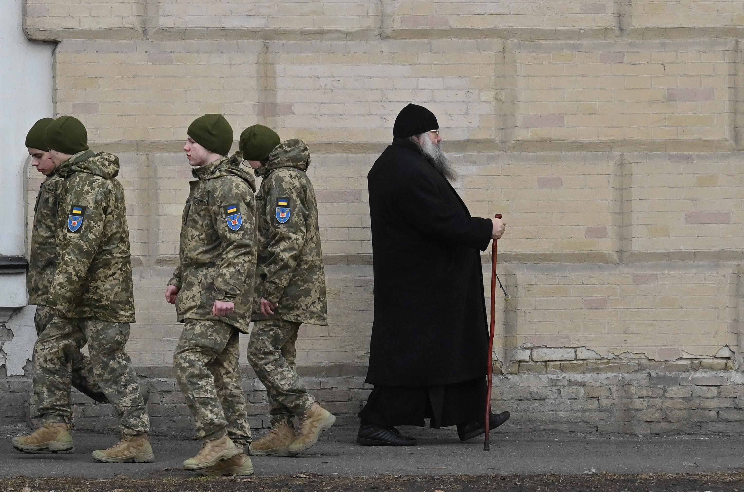 Prawosławny duchowny mija grupę ukraińskich żołnierzy na tle muru. Ukraińska Cerkiew Prawosławna.
