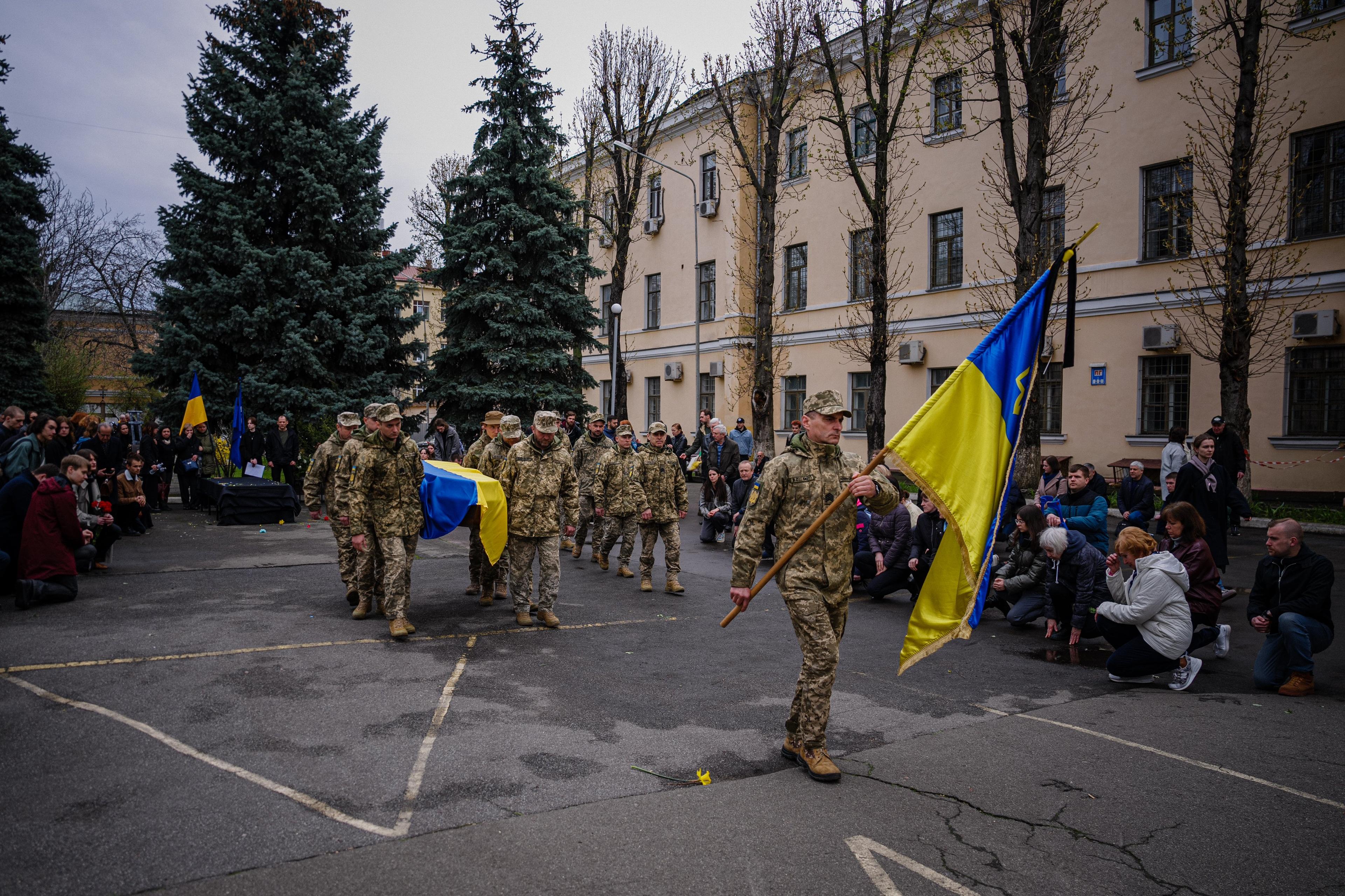 Żołnierze niosą trumnę przystrojoną ukraińską flagą, ludzie klękają