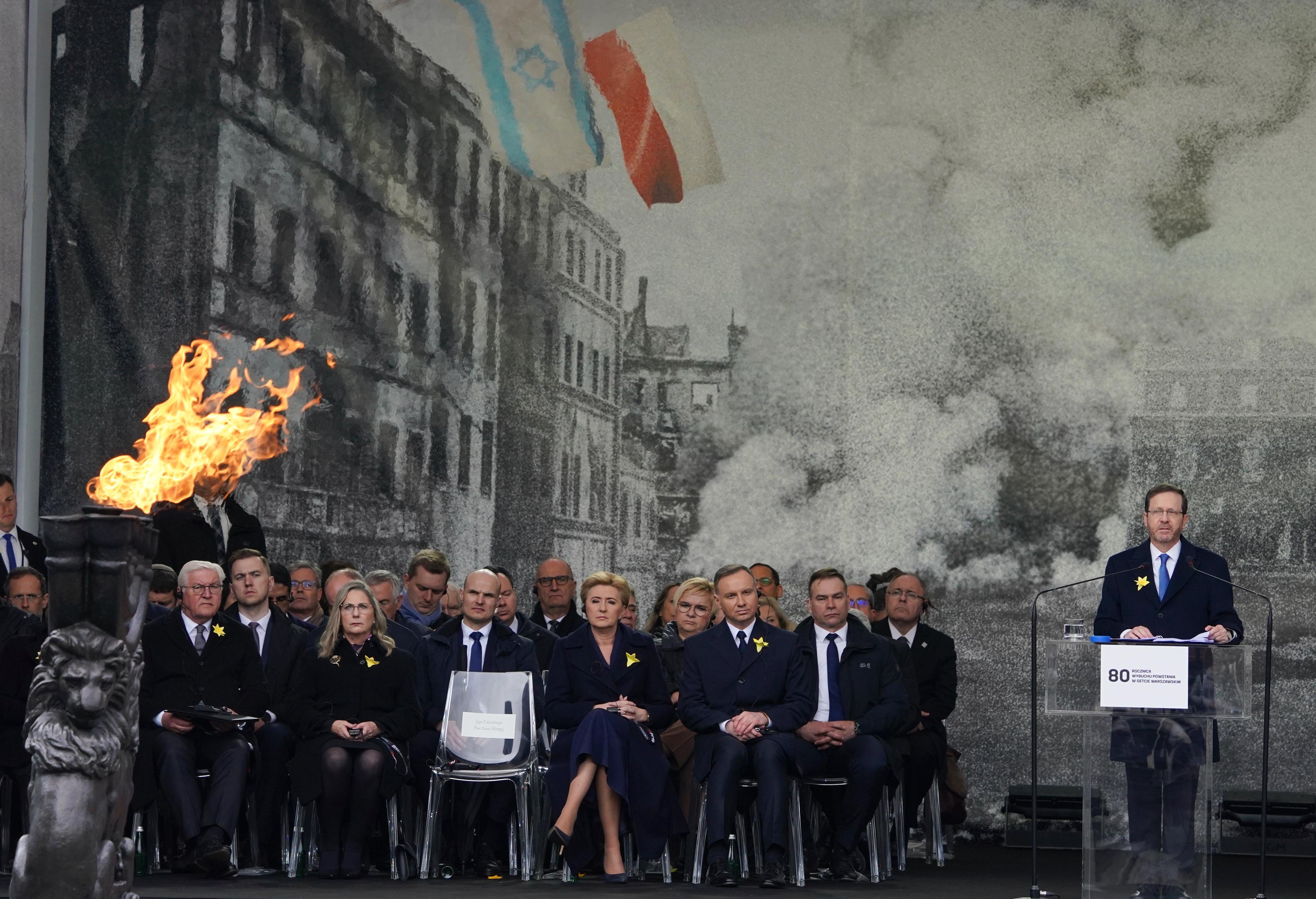 Oficjele siedzą na krzesłach na podeście. Za nimi duże czarno-białe zdjęcie płonącego warszawskiego getta. Na zdjęciu powiewają flagi Izraela i Polski