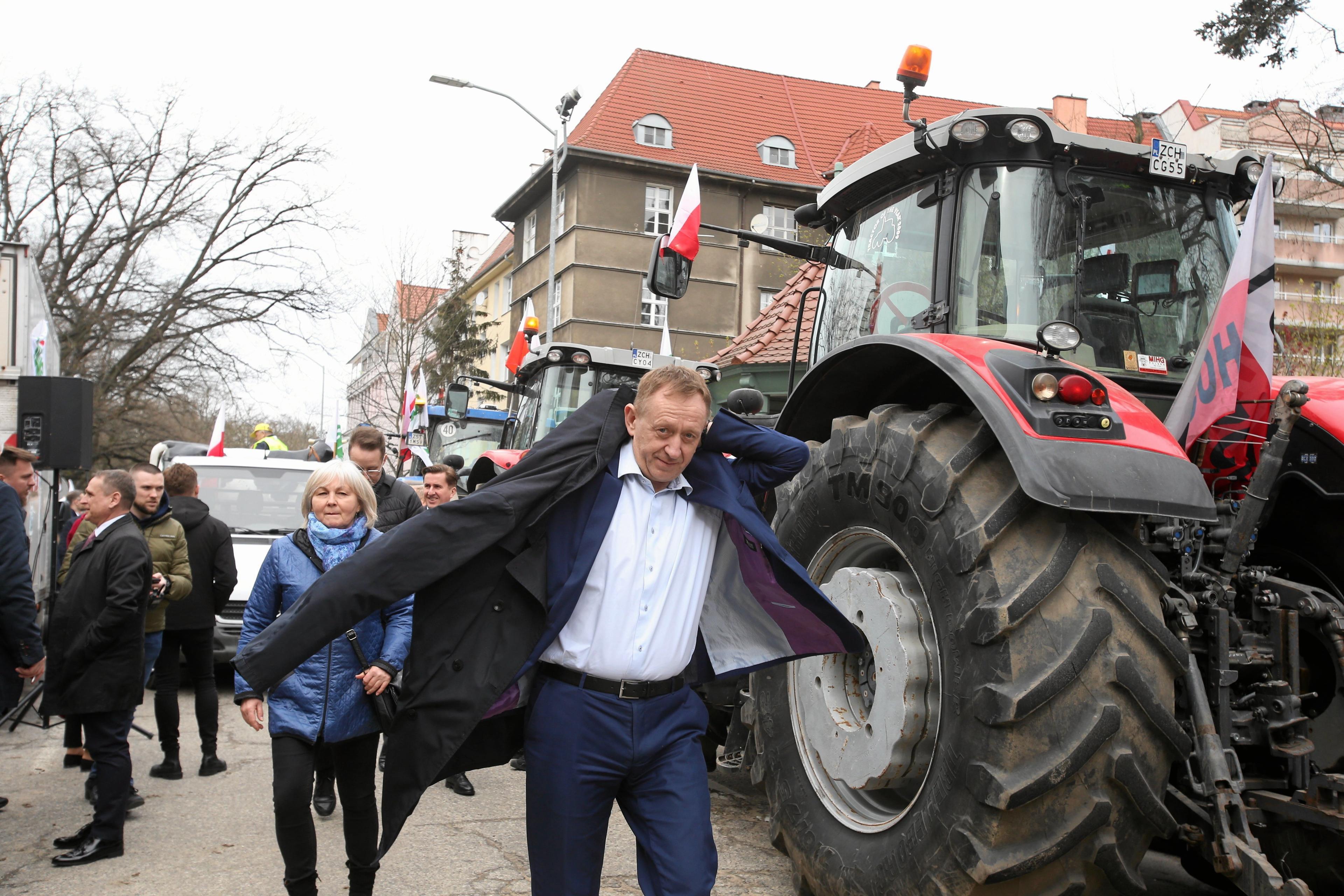 Minister Robert Telus po rozmowach z protestującymi rolnikami ubiera płaszcz i oddala się od rozmówców. W tle ciągnik rolniczy.