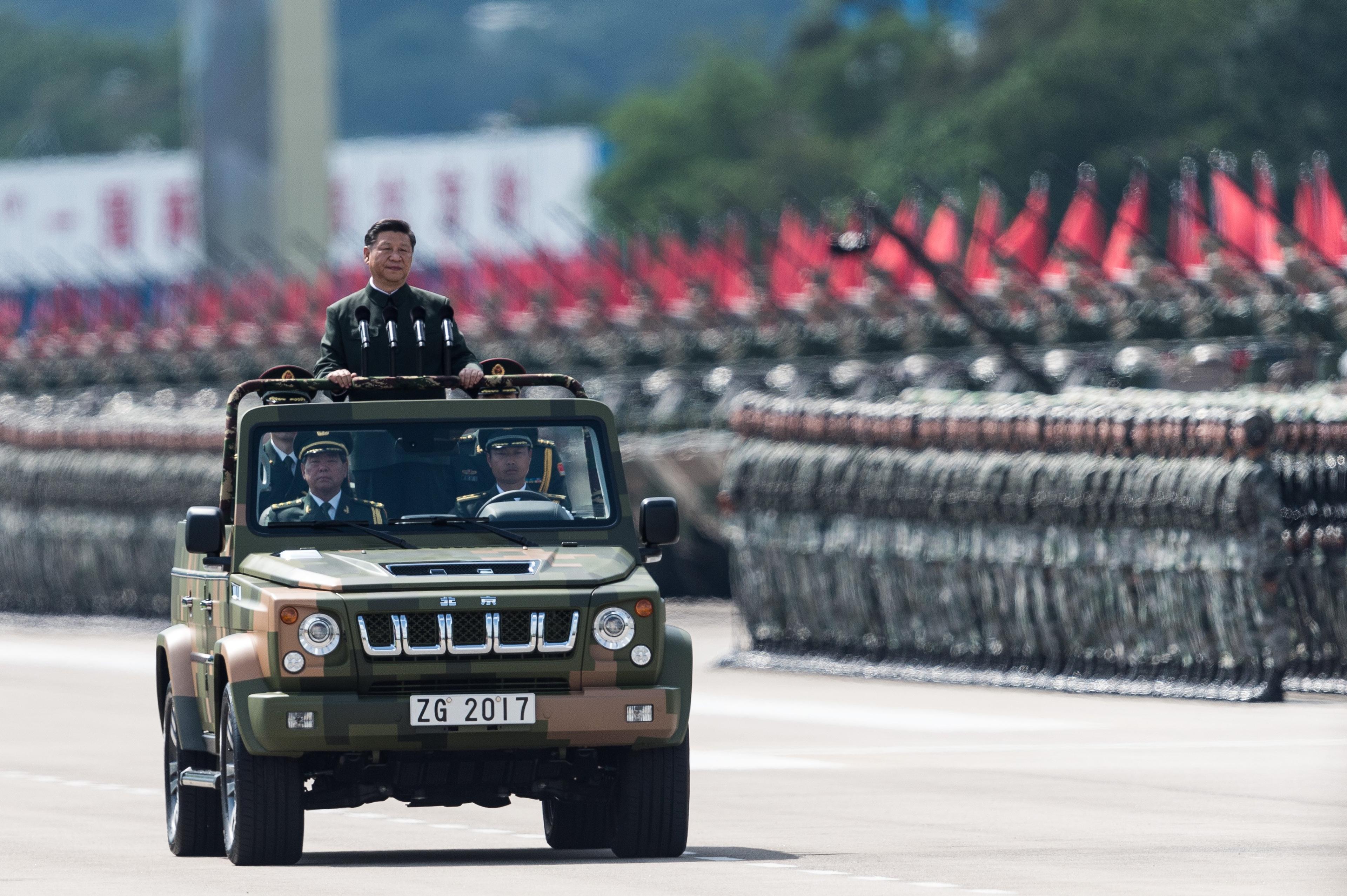Przewodniczący Chińskiej Republiki Ludowej Xi Jinping podczas defilady wojskowej. Chiński przywódca jest wieziony wojskowym samochodem tak, że wystaje ponad jego dach i może pozdrawiać żołnierzy. W tle równe zastępy wojska i uzbrojenia.