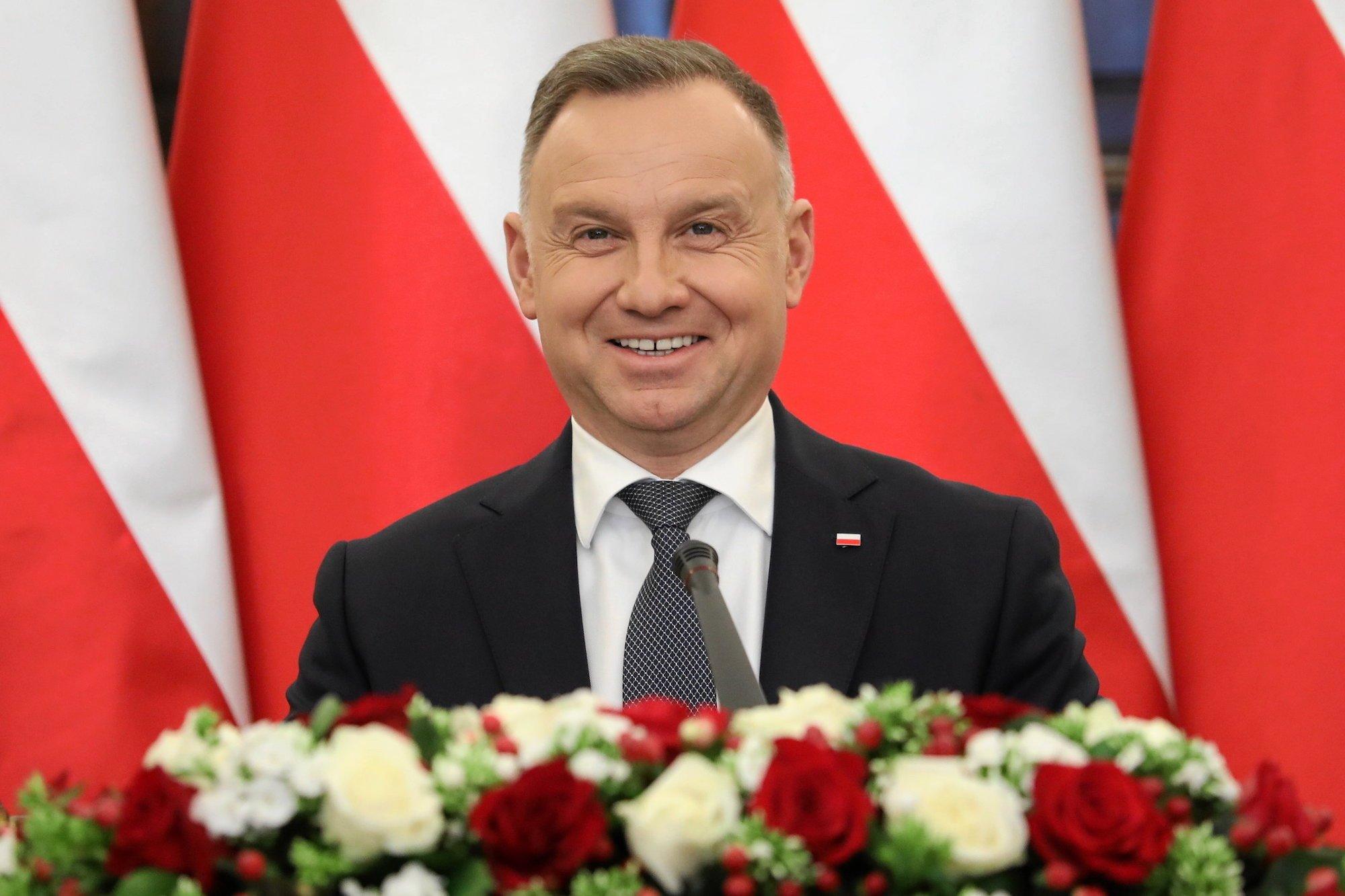 Szeroko uśmiechnięty prezydent Andrzej Duda na tle biało-czerwonych flag. Na pierwszym planie bukiet z białych i czerwonych róż.