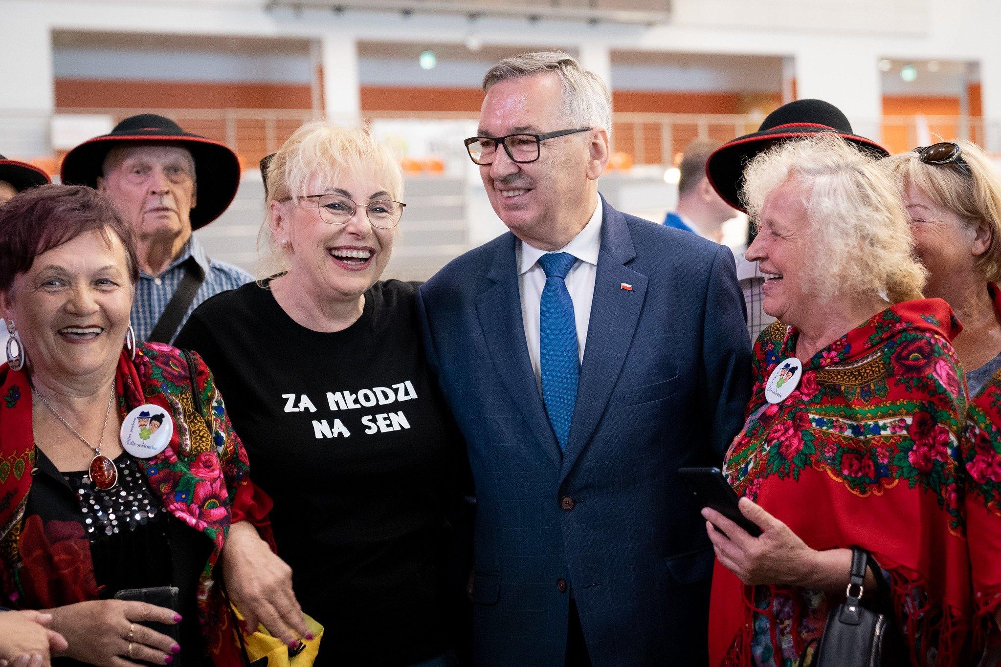 Stanisław Szwed w otoczeniu seniorów. Pani stojąca obok wiceministra ma na sobie czarną koszulkę z białym napisem "za młodzi na sen". Wszyscy szeroko się uśmiechają.