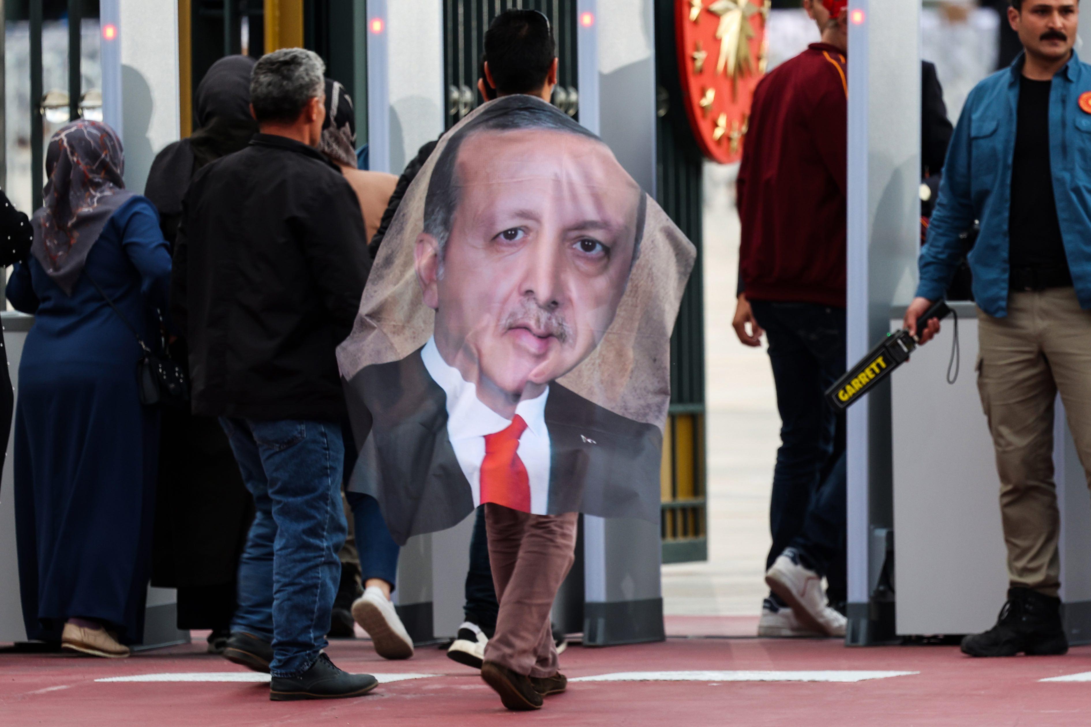 Wyborcy wchodzą prez bramkę bezpieczeństwa, jeden z nich ma na plecach płaszcz z fotografią Erdogana.